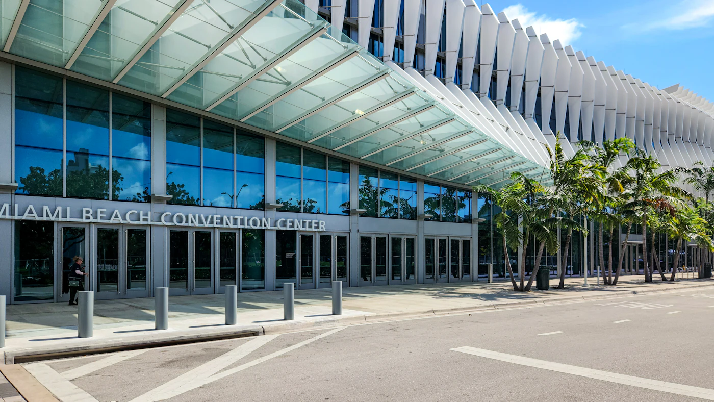 The glass facade and canopy of the Miami Beach Convention Center, lined with palm trees, on a sunny day
