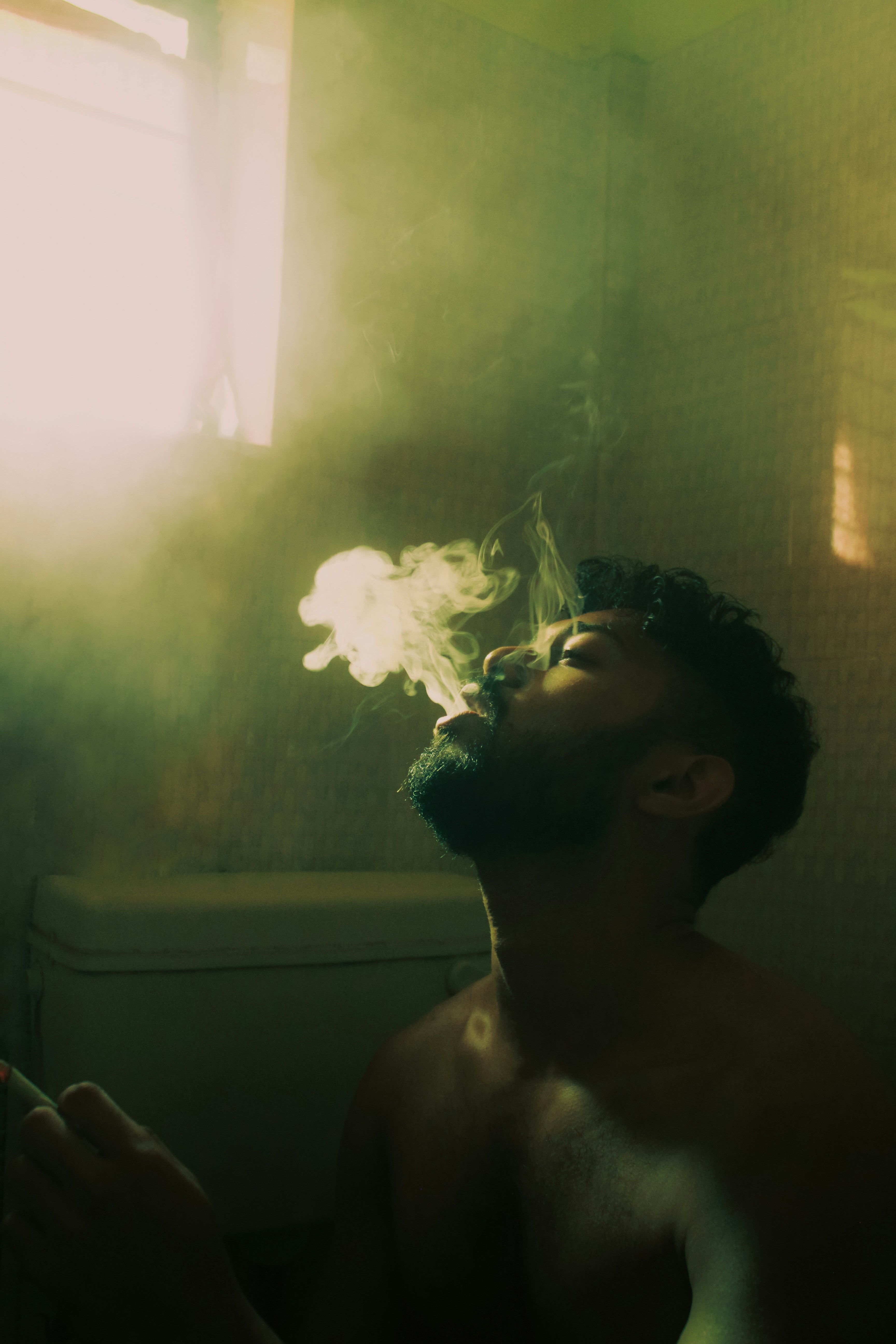 A shirtless man smoking a cigarette in front of a window