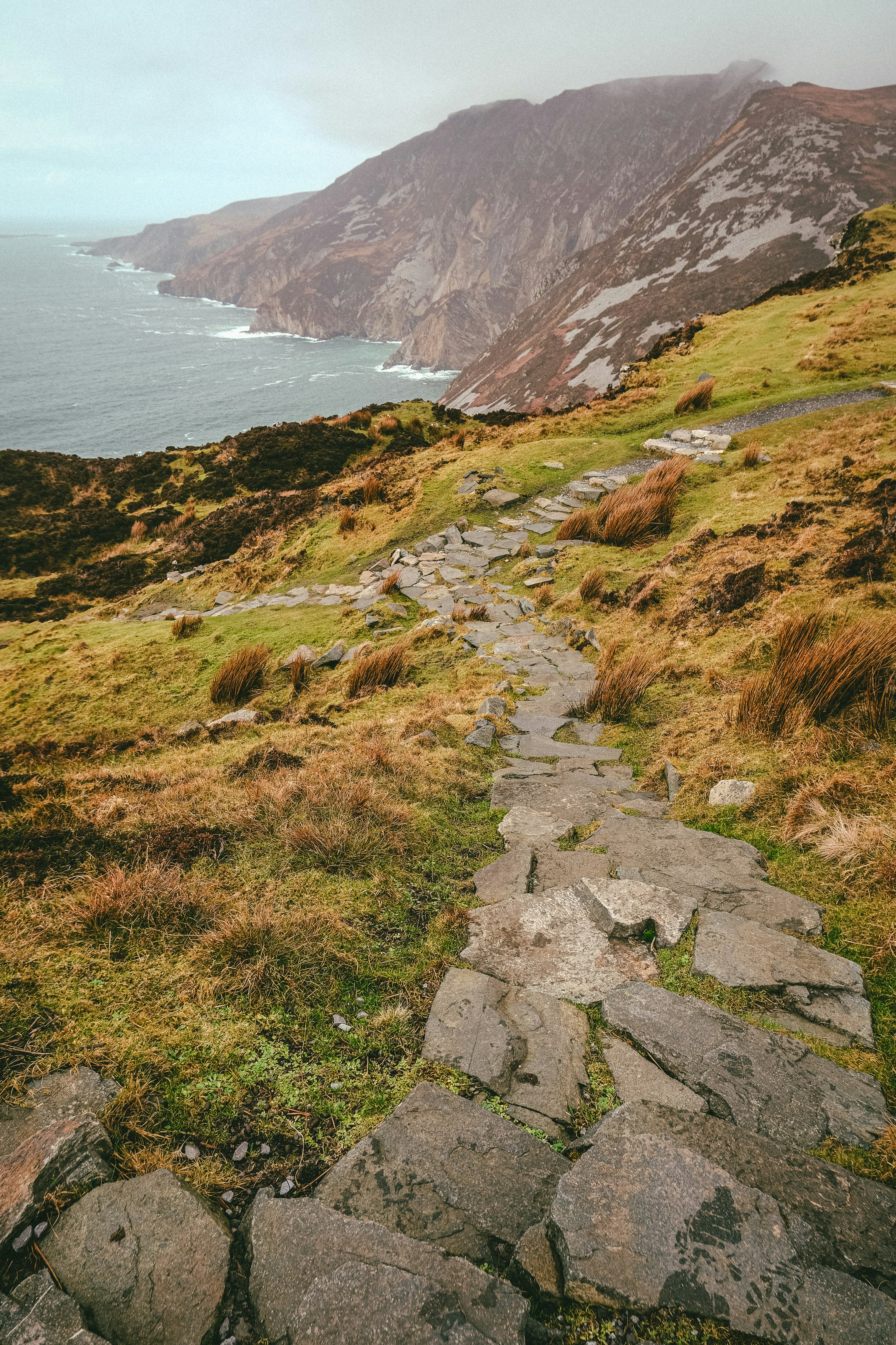 Stone path winds along a grassy clifftop toward the distant sea, with rugged cliffs in the background.
