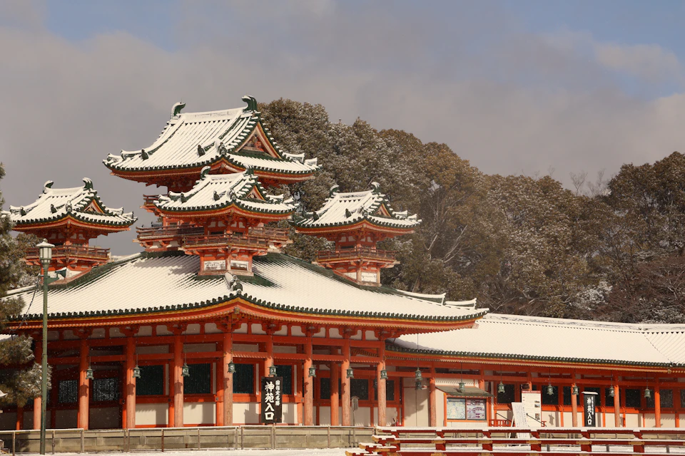The vermilion hall and green-tiled towers of Heian Shrine under a fresh layer of snow, with snow-dusted trees behind