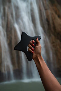 A hand holding a black object in front of a waterfall