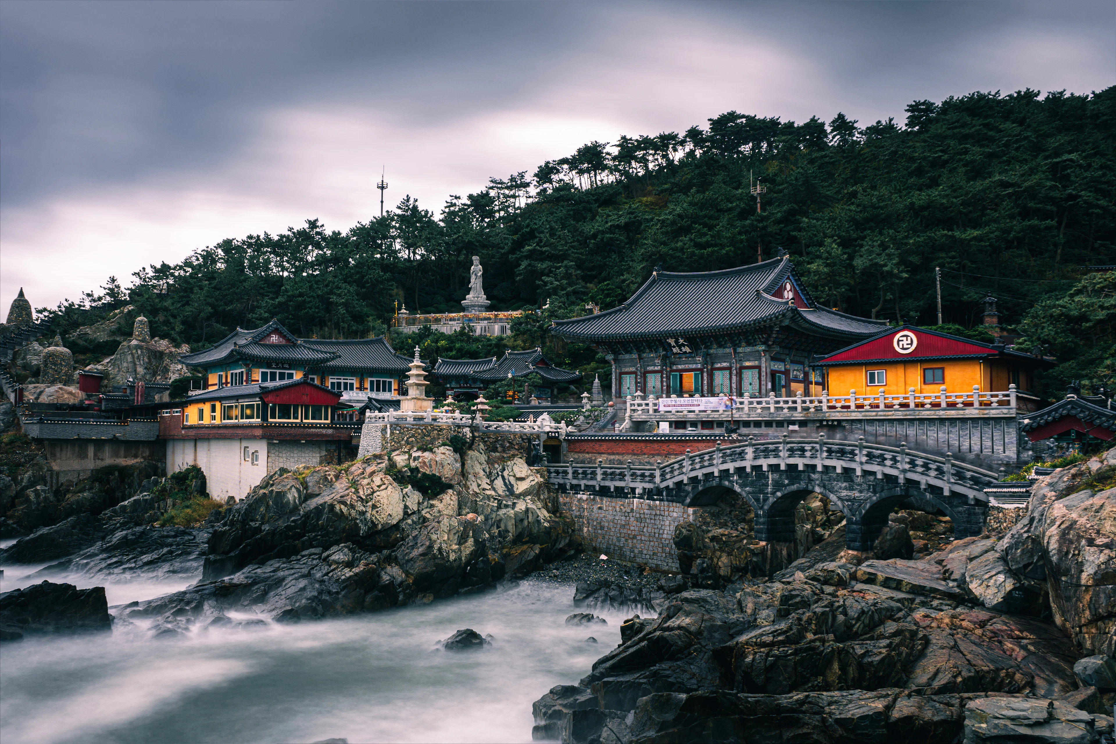 Haedong Yonggungsa temple in Busan built onto the black sea rocks, its tiled roofs and yellow and red halls stacked below a wooded headland, the surf blurred to mist by a long exposure under a heavy grey sky.