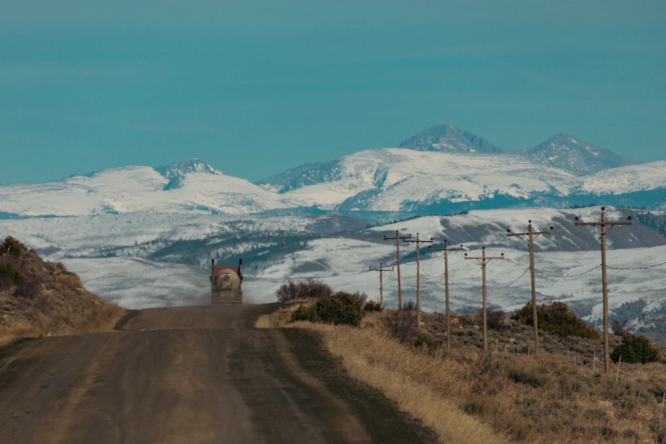The image depicts a dirt road winding through a mountainous landscape, with snow-capped mountains rising against a clear blue sky. A large truck is driving on the right side of the road, leaving behind a trail of dust that contrasts with the pristine white snow covering the mountain tops and valley below. The scene conveys a sense of solitude and tranquility as the truck moves through the vast expanse of nature.

The mountains in the background are covered in snow, their peaks reaching towards the sky, while the valley is blanketed by dry grass that sways gently with the wind. The road itself is a dirt path, its surface marked by tire tracks and dust from the truck's journey.