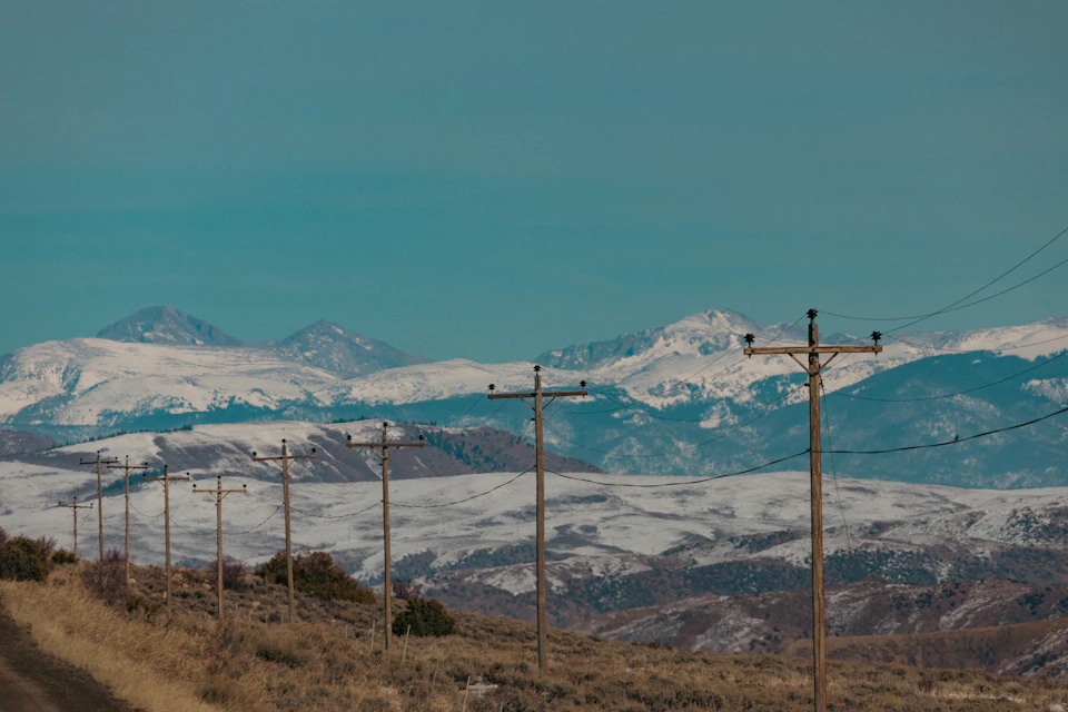 The image depicts a serene winter landscape with snow-capped mountains and power lines running across the frame, creating a harmonious blend of natural beauty and human infrastructure. The perspective is from ground level, looking up at the majestic peaks in the distance. The sky above is a clear blue, providing a striking contrast to the white snow on the mountain tops.

The power lines are visible as they stretch across the image, with some closer to the foreground and others further away. The mountains rise majestically in the background, their peaks dusted with snow that glistens under the sunlight.