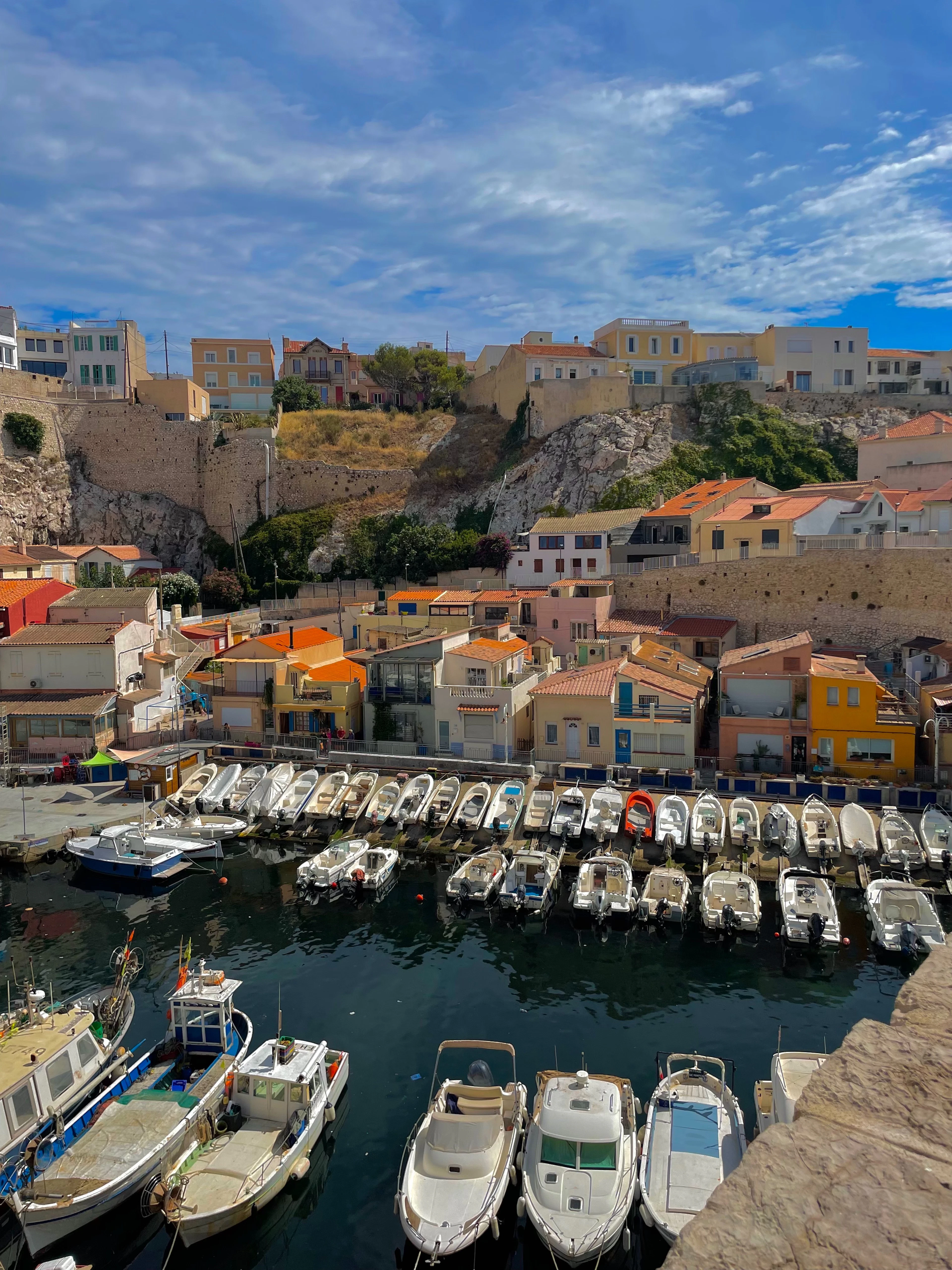 The tiny harbour of Vallon des Auffes in Marseille packed rail to rail with small white boats, the pastel yellow, orange and pink fishermen's houses stacked around the water and the stone viaduct wall rising behind them under a blue sky.