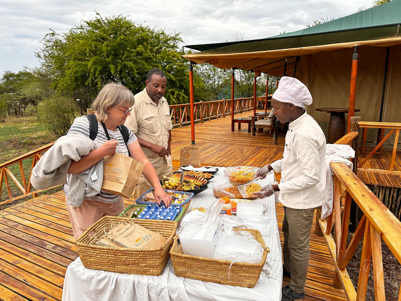 Guests serving themselves from a safari camp dining table on a deck, a tented camp behind