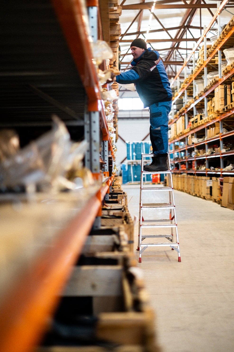 Warehouse worker on ladder accessing high shelf in tall metal storage rack