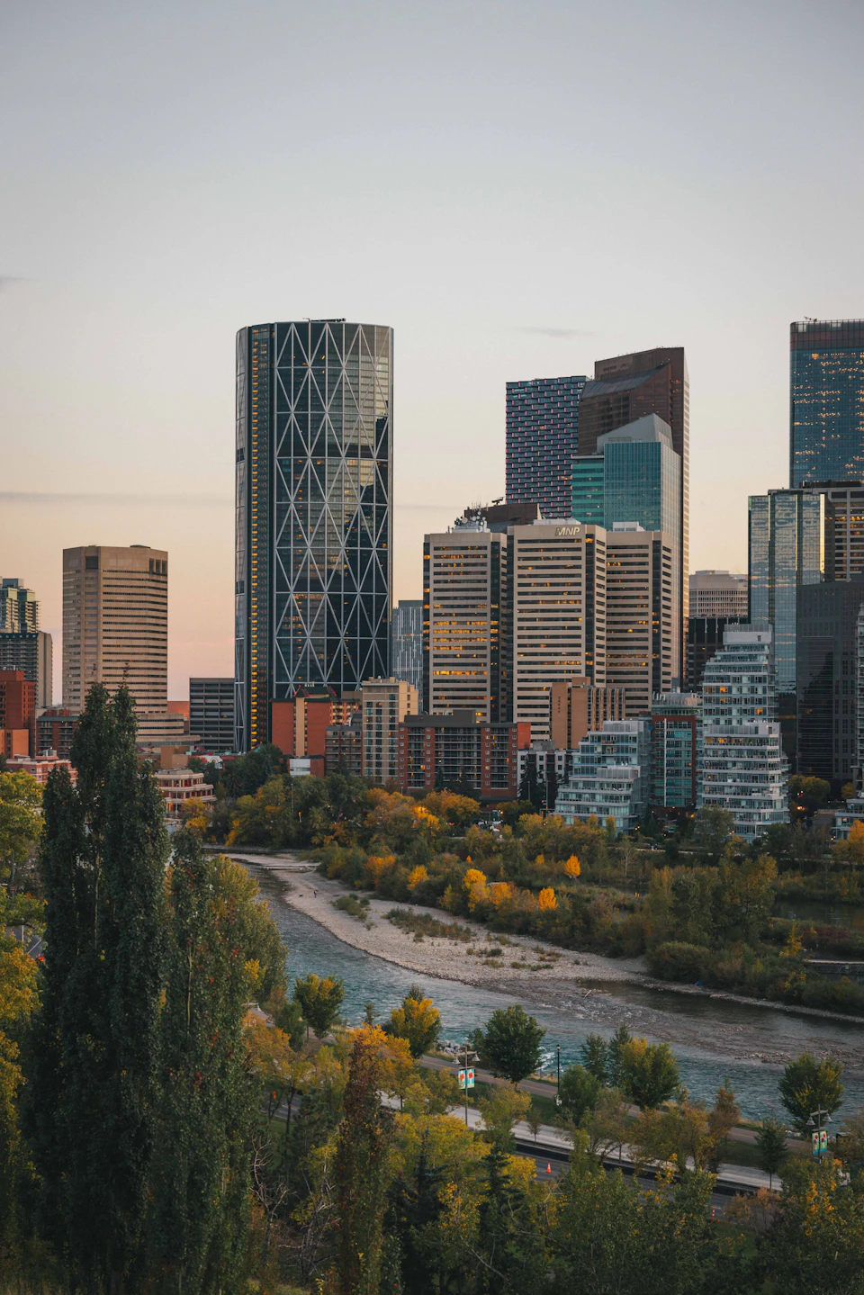 The image presents a panoramic view of the downtown core of a major city, with the skyline filled with tall buildings that stretch towards the sky. The tallest building is a modern skyscraper with a unique curved facade and a glass exterior that reflects the surrounding environment. In the foreground, there are trees lining the riverbank, adding a touch of nature to the urban landscape. The colors in the image are predominantly gray and brown, reflecting the city's architectural style and the natural elements present along the river.