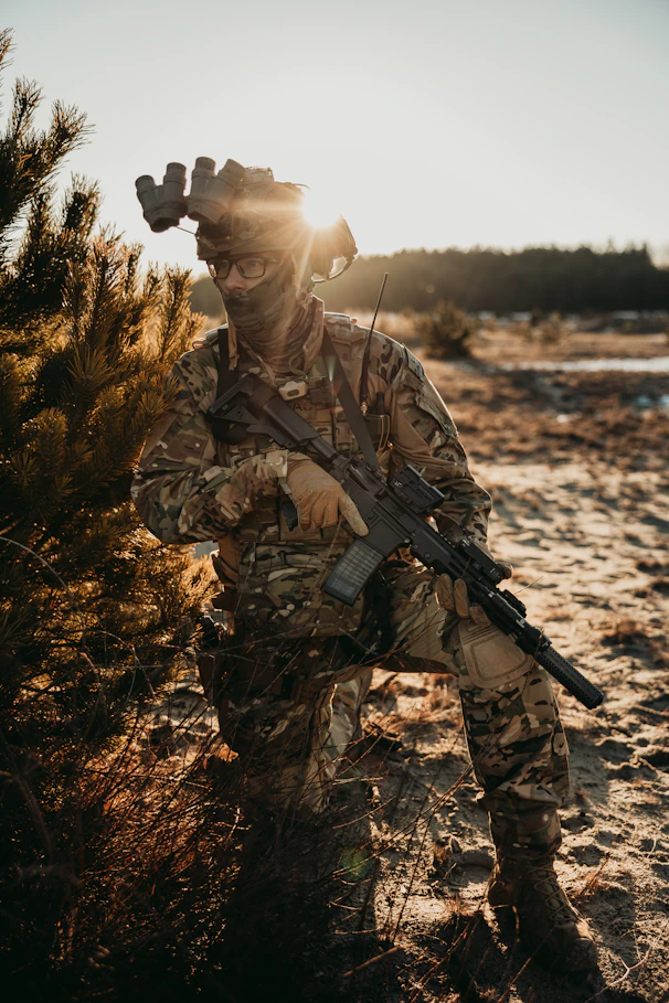 A soldier kneels in camouflage at sunset.