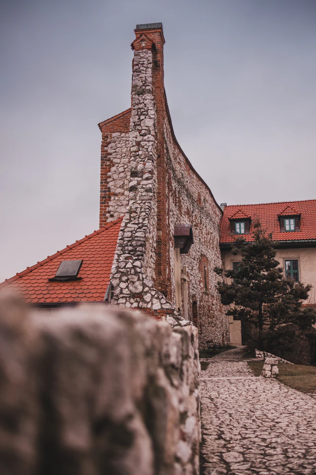 A stone and brick monastery building with a tall chimney and red tile roofs beside a cobblestone path and stone wall.