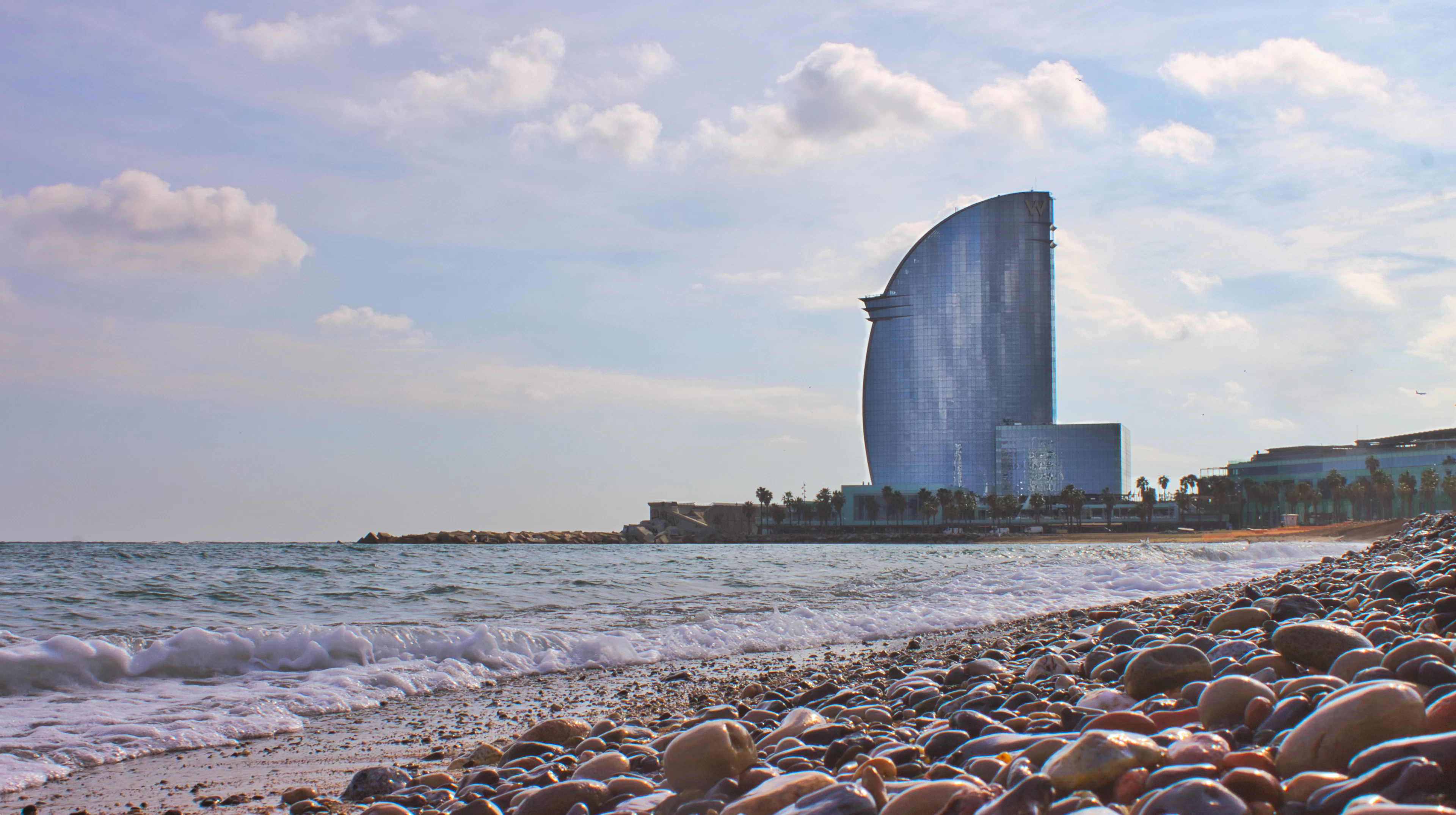 The sail-shaped W Barcelona hotel rising at the end of the pebbled Barceloneta shoreline, waves breaking in the foreground