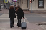 Two people walk across a crosswalk with luggage.