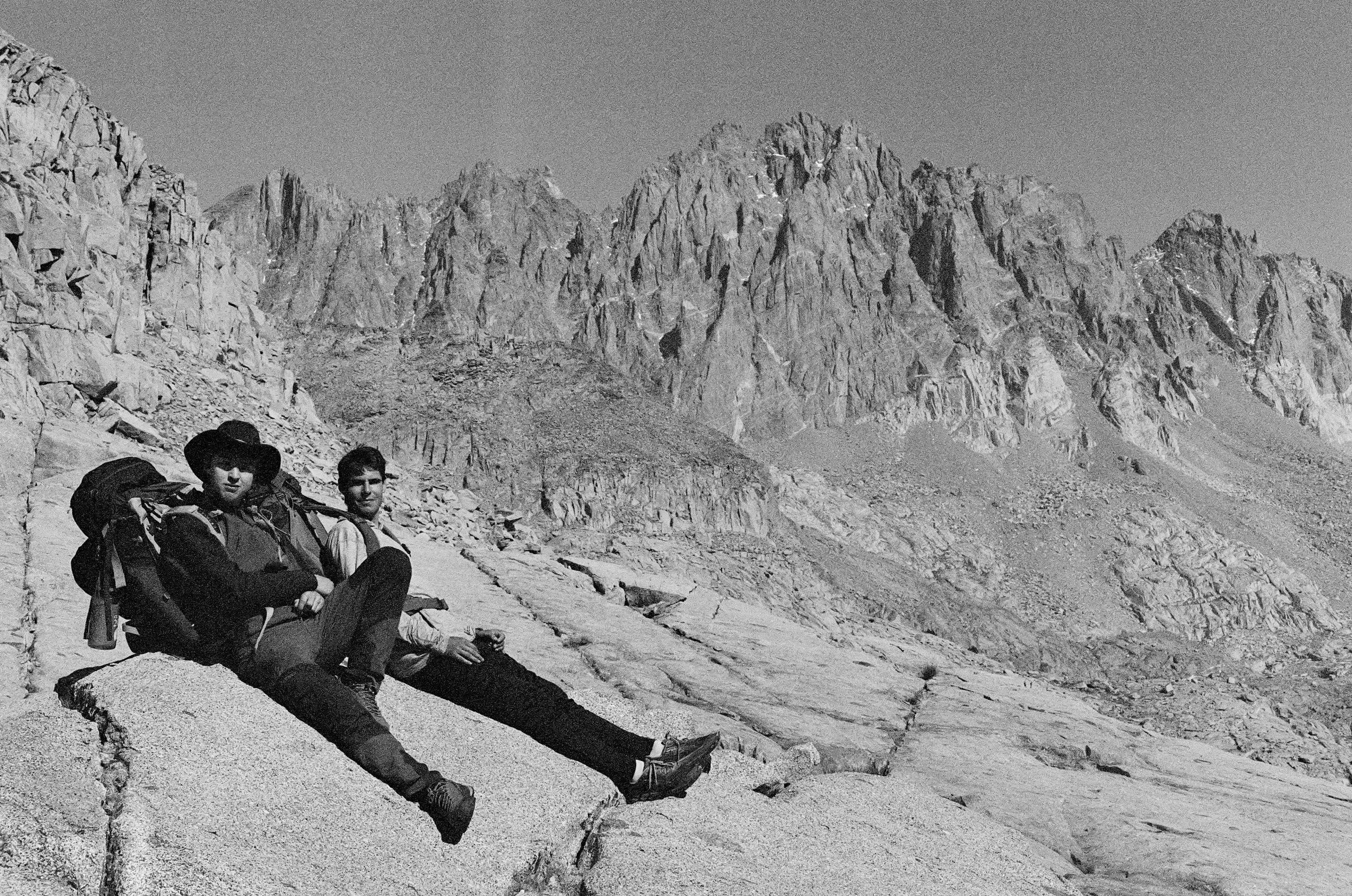 Two hikers resting on a rock ledge with jagged mountain peaks in the background