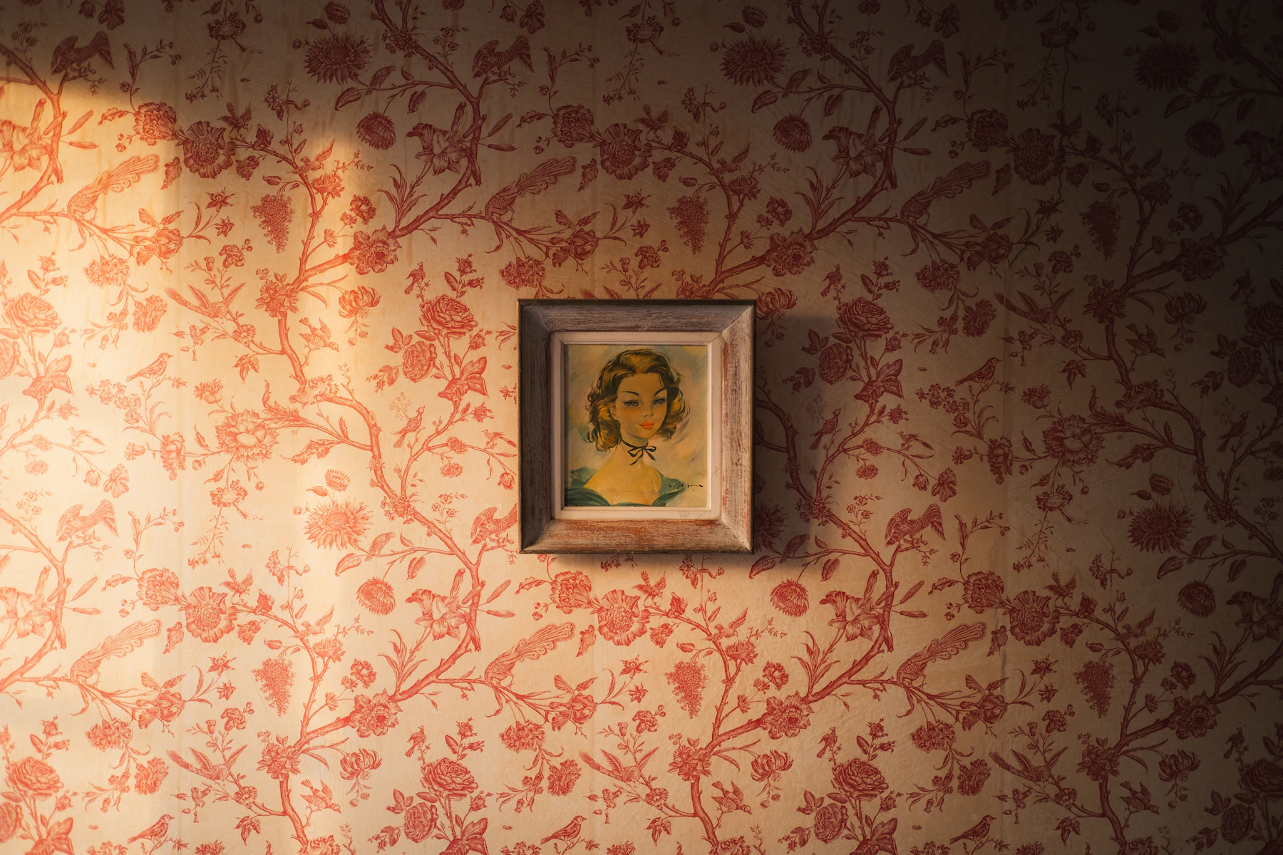 A small framed portrait hanging on a wall papered in a red floral pattern