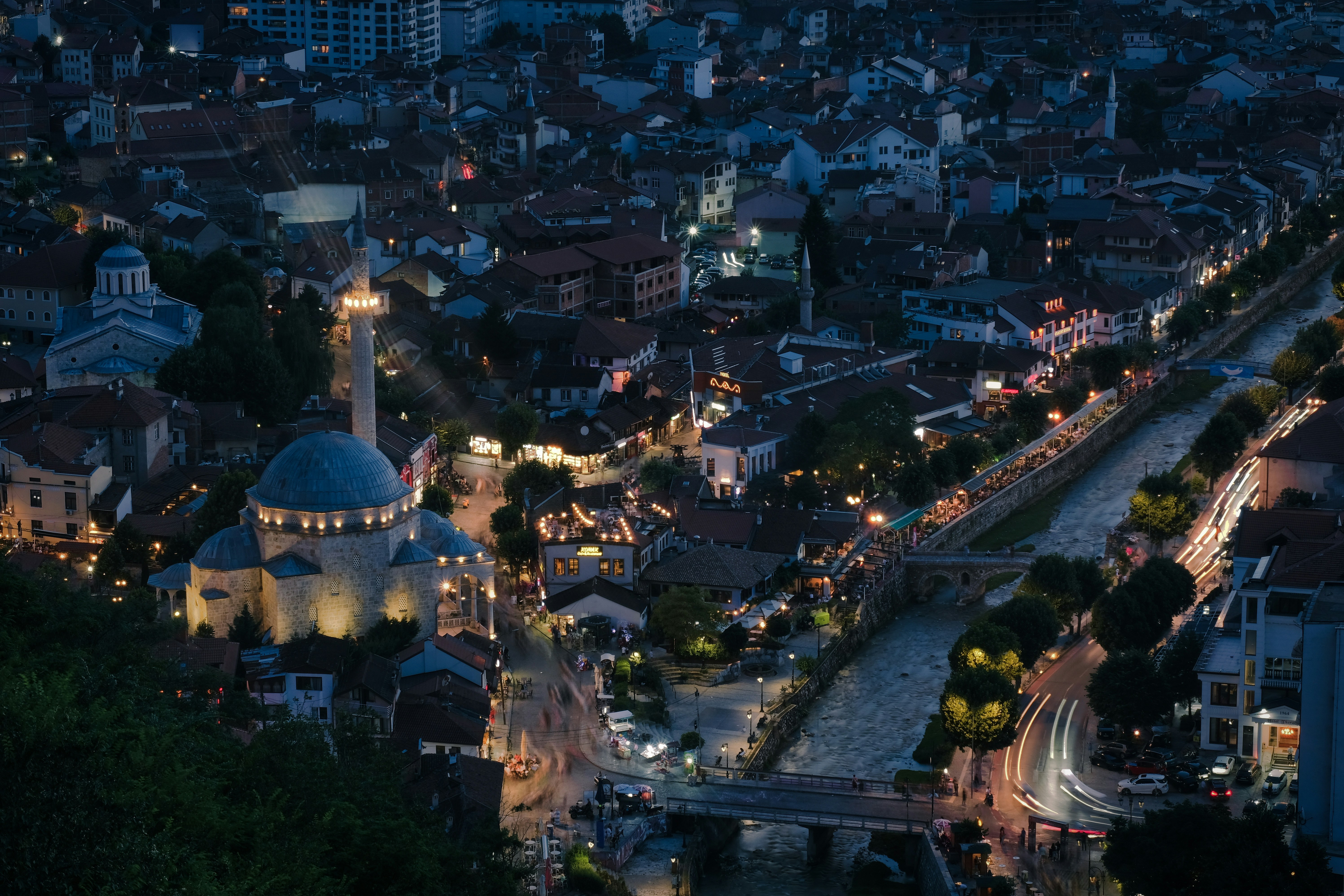 Prizren, a city where light and history intertwine. As dusk falls, the warm glow of streetlights and the illuminated mosque breathe life into the historic heart of the city. The winding river, stone bridges, and vibrant streets tell a story of culture, tradition, and timeless beauty. A perfect blend of past and present under the evening sky.