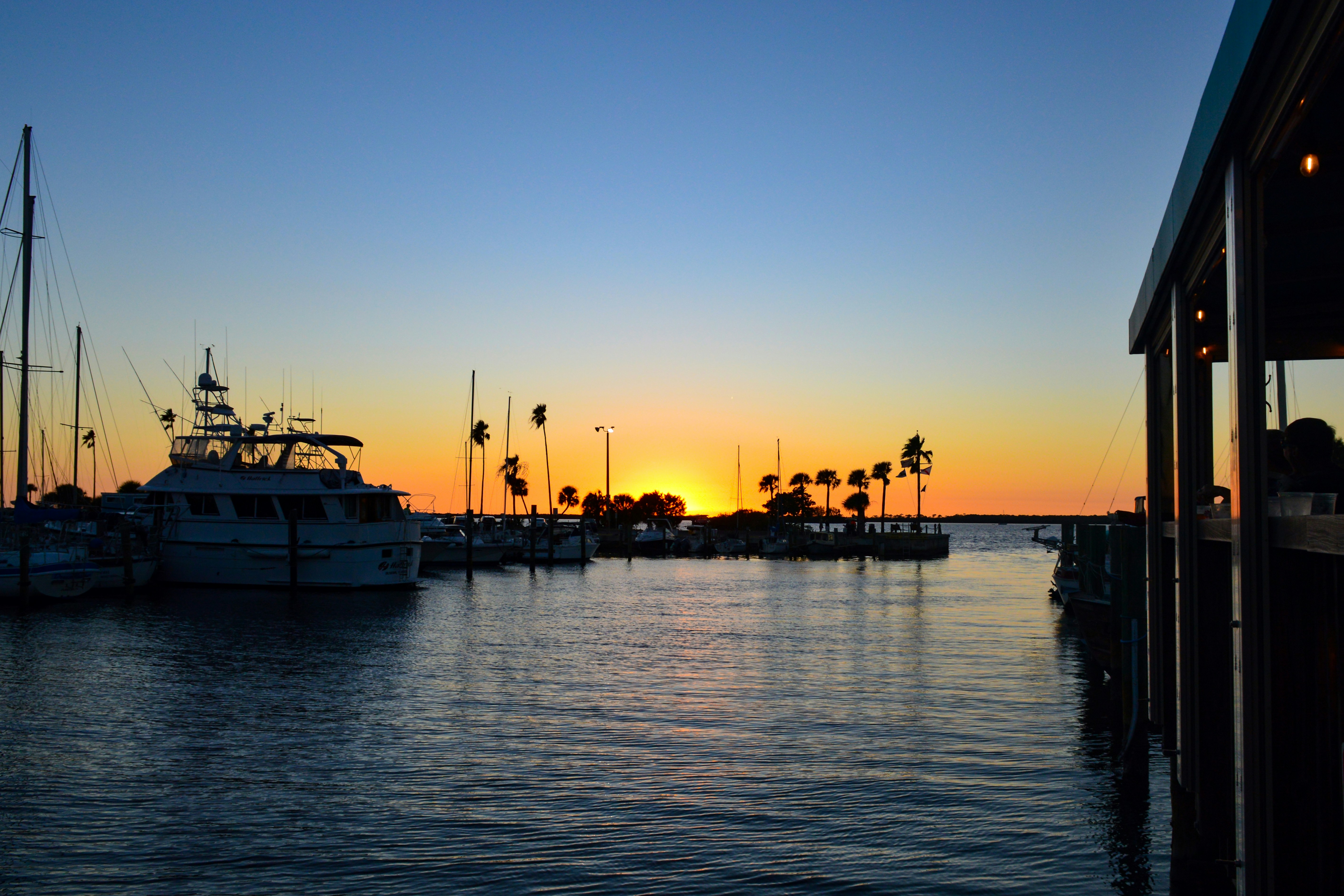 A peaceful marina at sunset, with sailboats and yachts anchored in calm waters. The sun sets on the horizon, casting a golden glow that fades into soft blue as evening approaches. Silhouettes of palm trees line the shore.