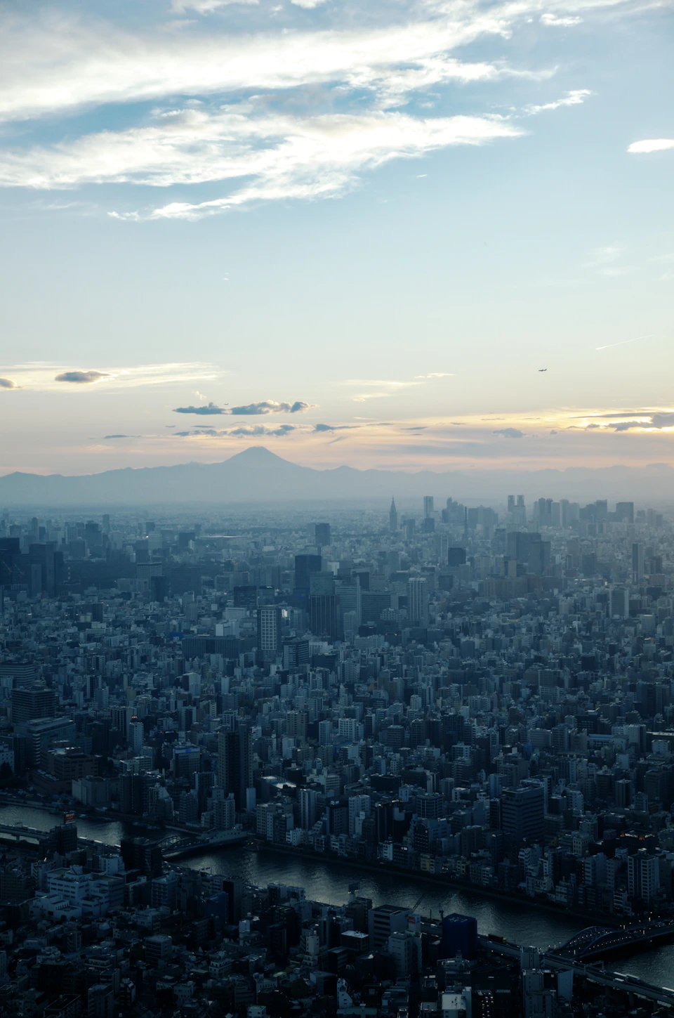 The image presents a panoramic view of a cityscape, captured from an elevated perspective. The city is densely populated with buildings of varying heights and designs, creating a sense of depth and scale. A river flows through the heart of the city, adding a touch of natural beauty to the urban landscape. The sky above is painted in shades of blue, dotted with fluffy white clouds that cast soft shadows on the city below.
