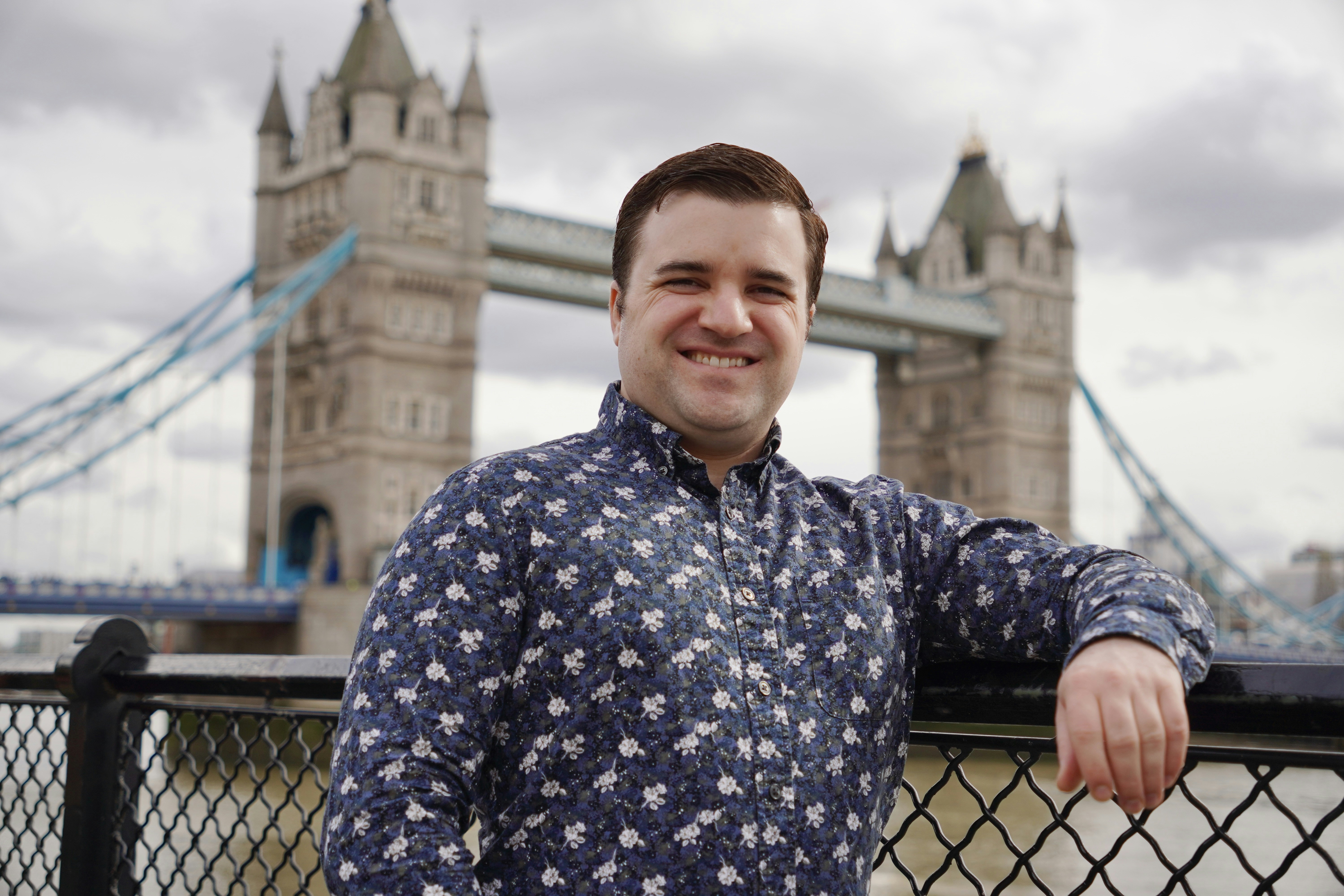 Photographer Brett Wharton poses with a smile in front of the iconic Tower Bridge in London, capturing a moment of joy and exploration in the historic city. The overcast sky and the soft lighting add a natural, candid feel to the portrait, with the famous landmark serving as a perfect backdrop.
