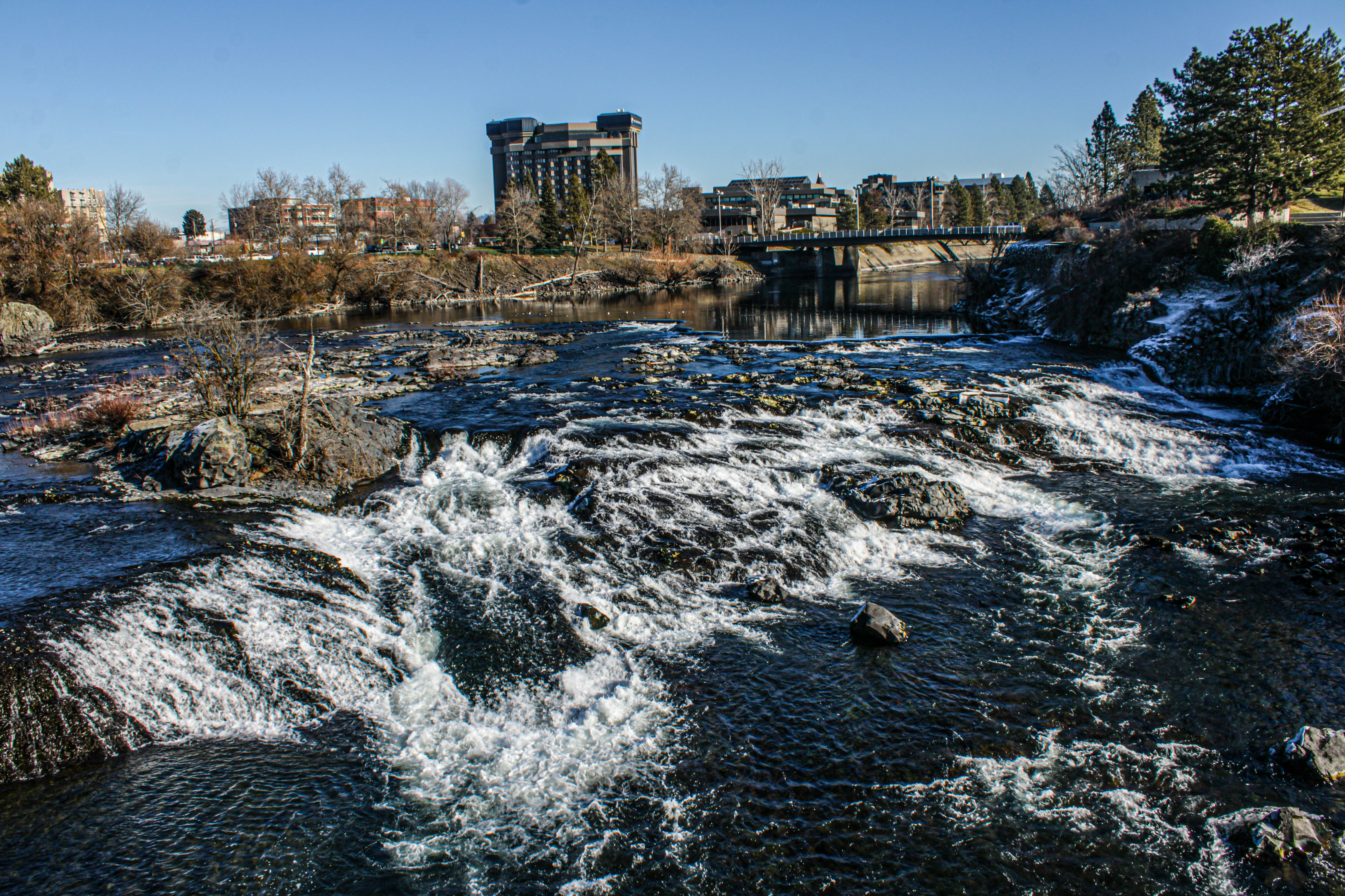 Water rushes over rocks, a cityscape in the background.