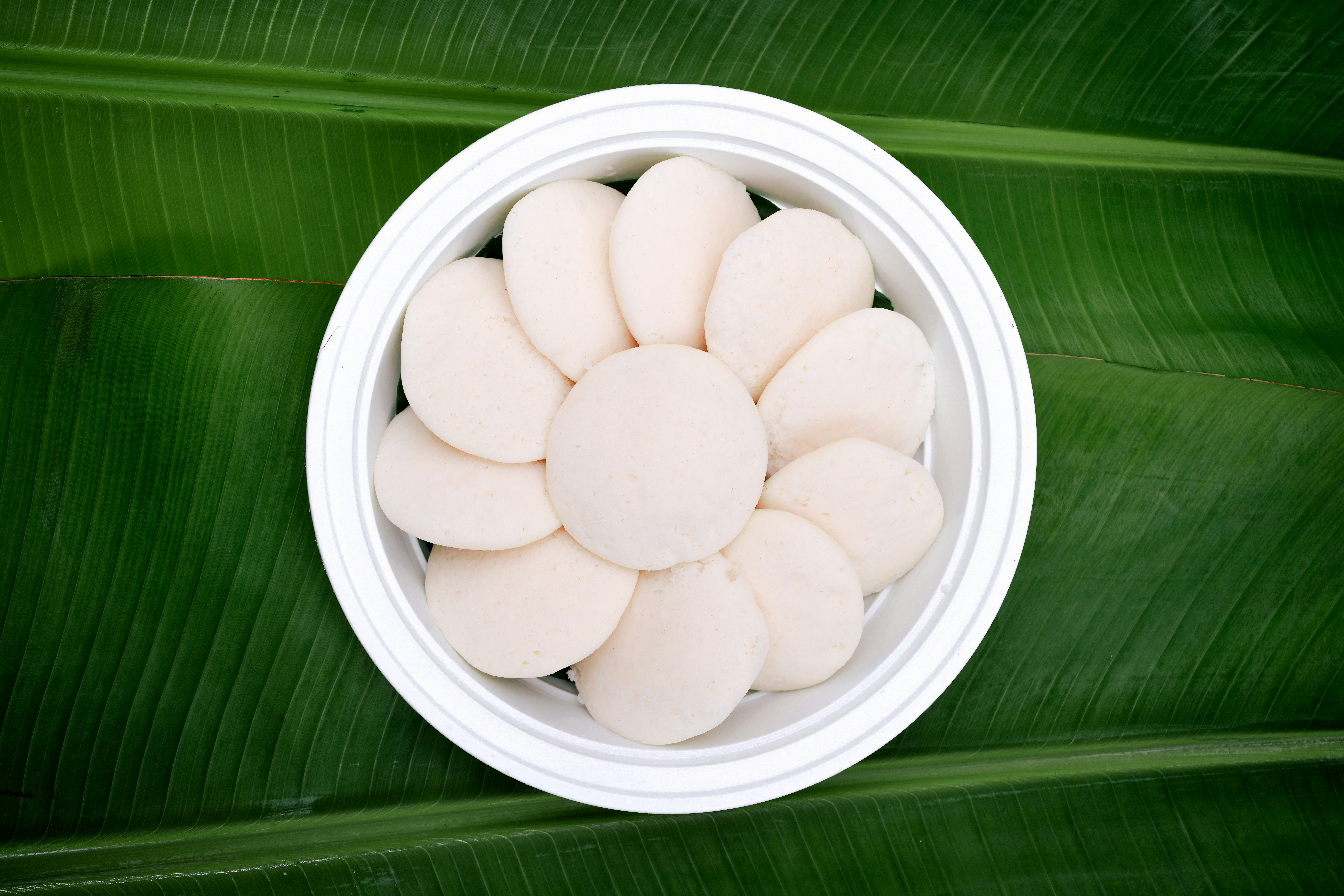 Steamed idli arranged in a circle in a white bowl on a green leaf