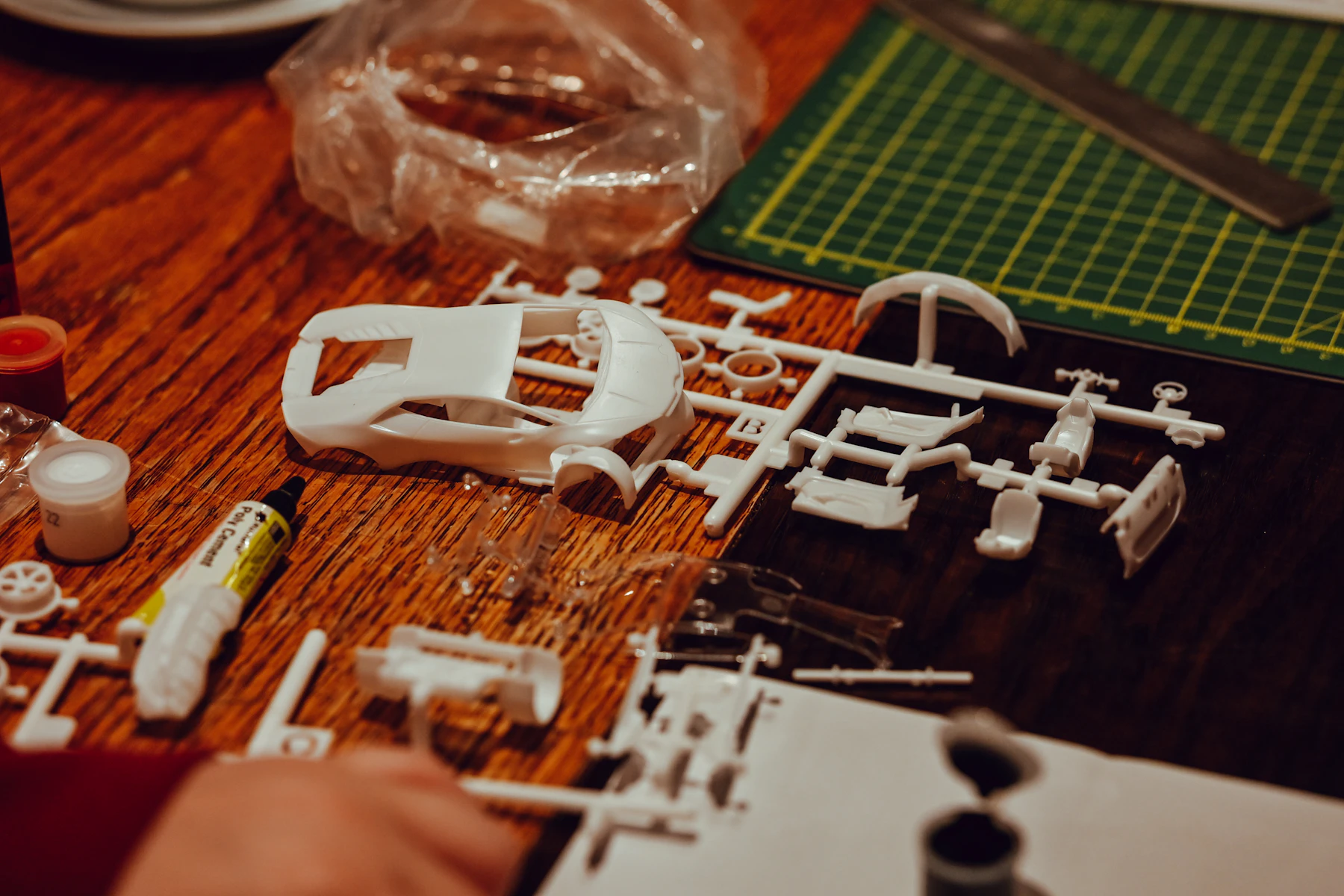 An unpainted white model car body and loose parts on a wooden table, with cement, paint pots and a green cutting mat behind