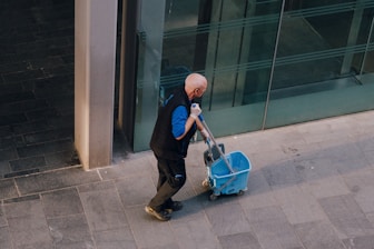 A man pulls a cleaning cart near a building.