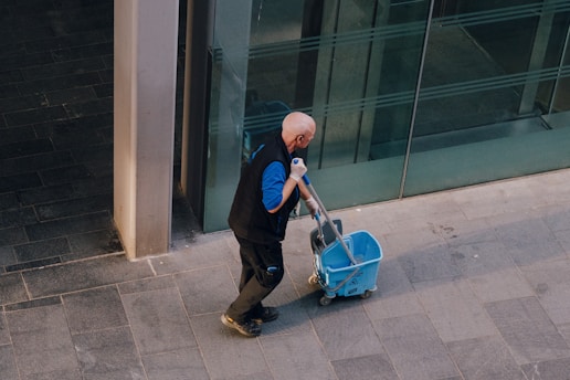 A man pulls a cleaning cart near a building.