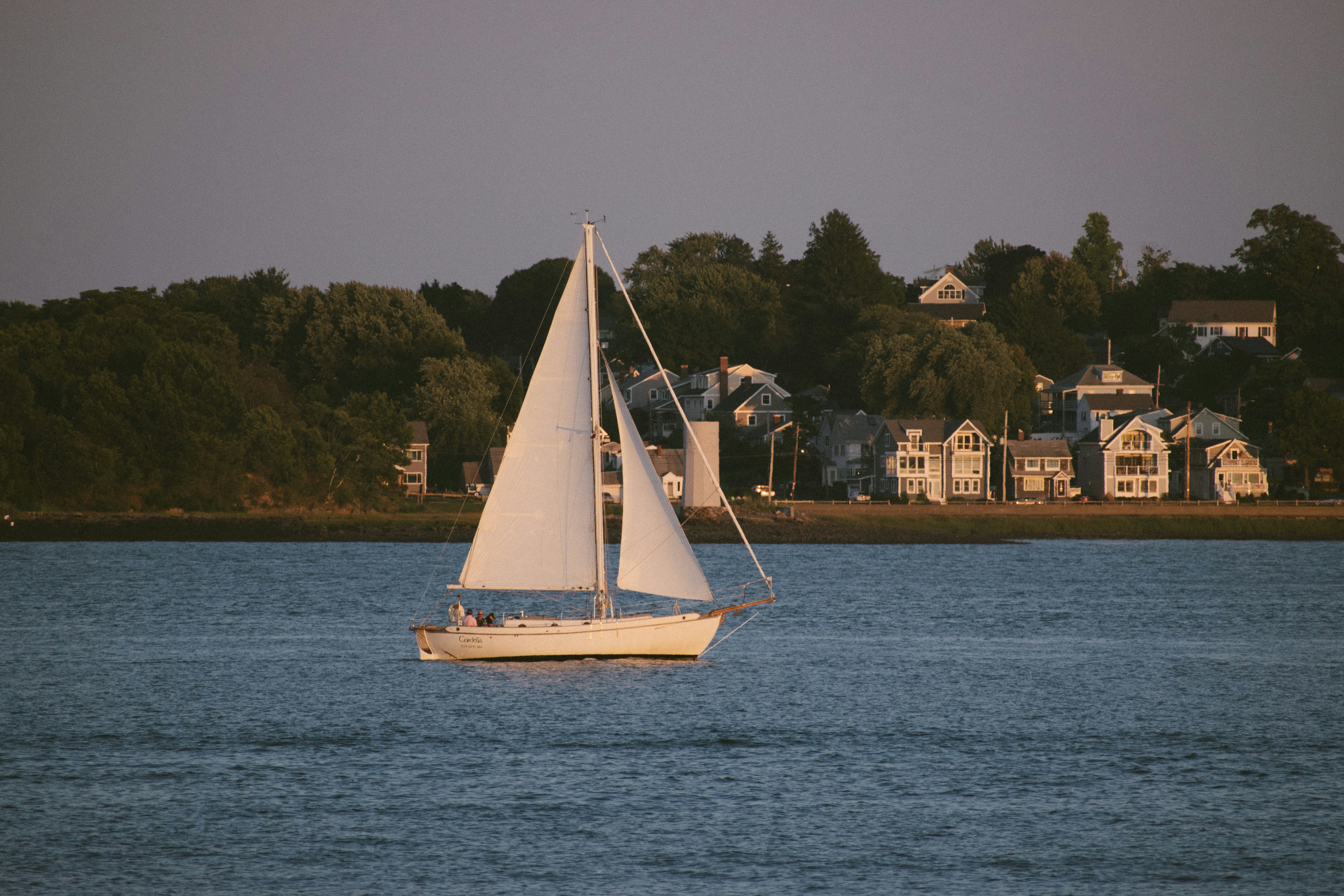 Lone daysailer sailboat beating out of a UK harbour entrance under soft evening light while the coastline fades behind.