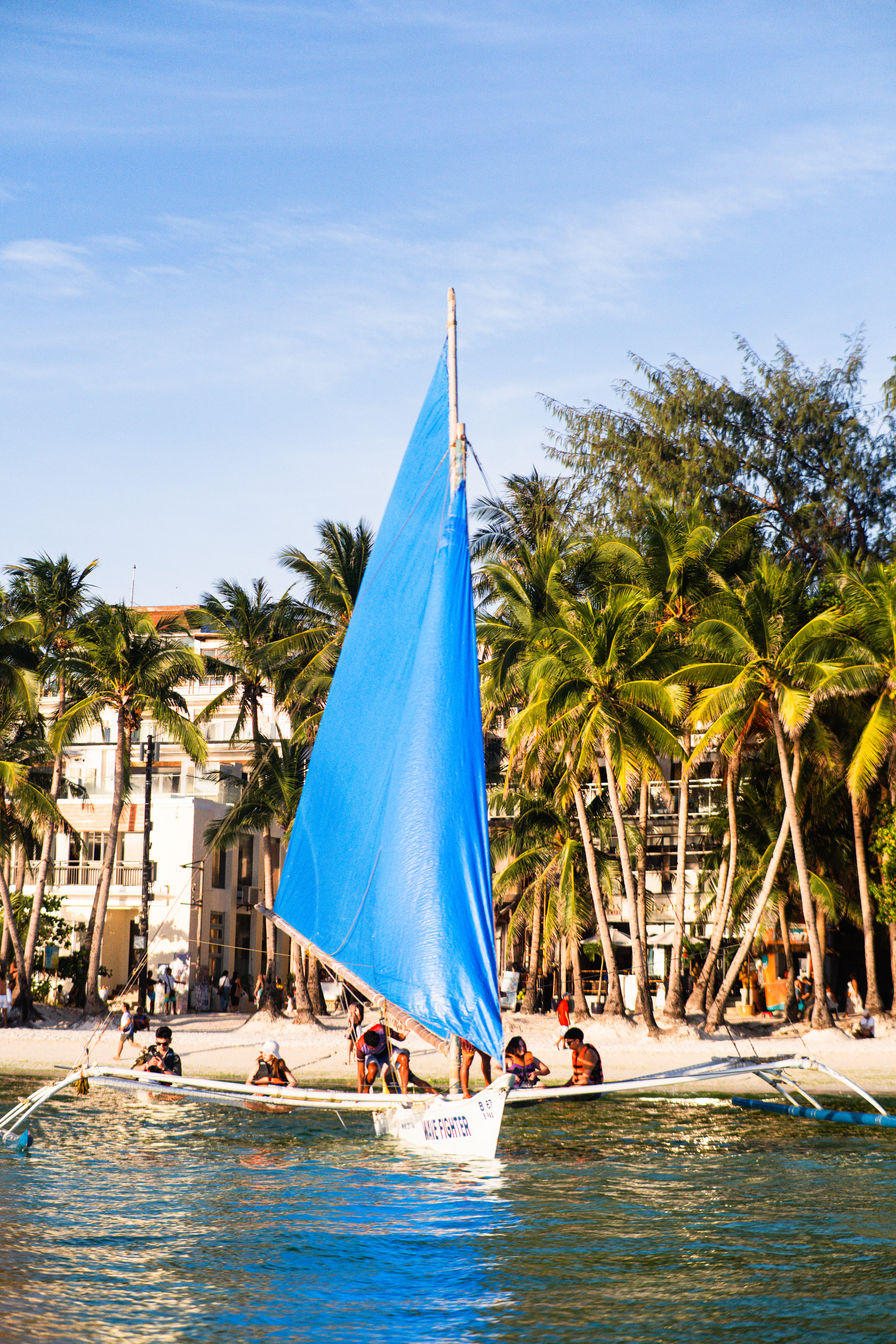 A paraw outrigger sailboat with a bright blue sail drawn up on the shallows off Boracay's White Beach, with leaning coconut palms and beachfront resort buildings behind