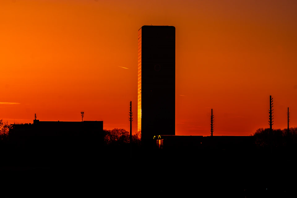 The image depicts a tall, rectangular building with a pointed top and a flat base, standing against an orange sky. The building is illuminated by warm light from below, creating a silhouette effect that contrasts with the vibrant colors of the sunset. In the foreground, there are trees and power lines visible in the distance, adding depth to the scene.

The image is taken from a low angle, making the building appear even more imposing against the backdrop of the sky. The warm orange hues of the setting sun cast long shadows on the ground, enhancing the dramatic effect of the photograph.