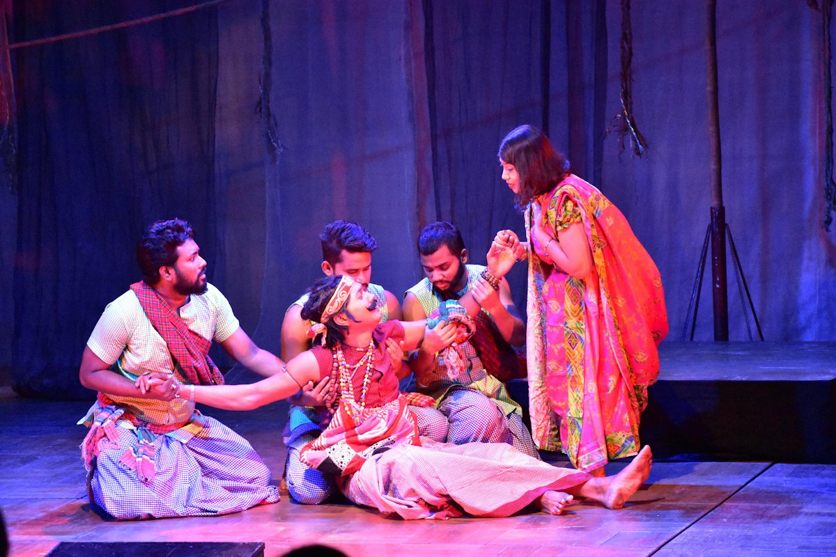 A cast in colorful costumes gathered around a figure lying on a blue-lit stage