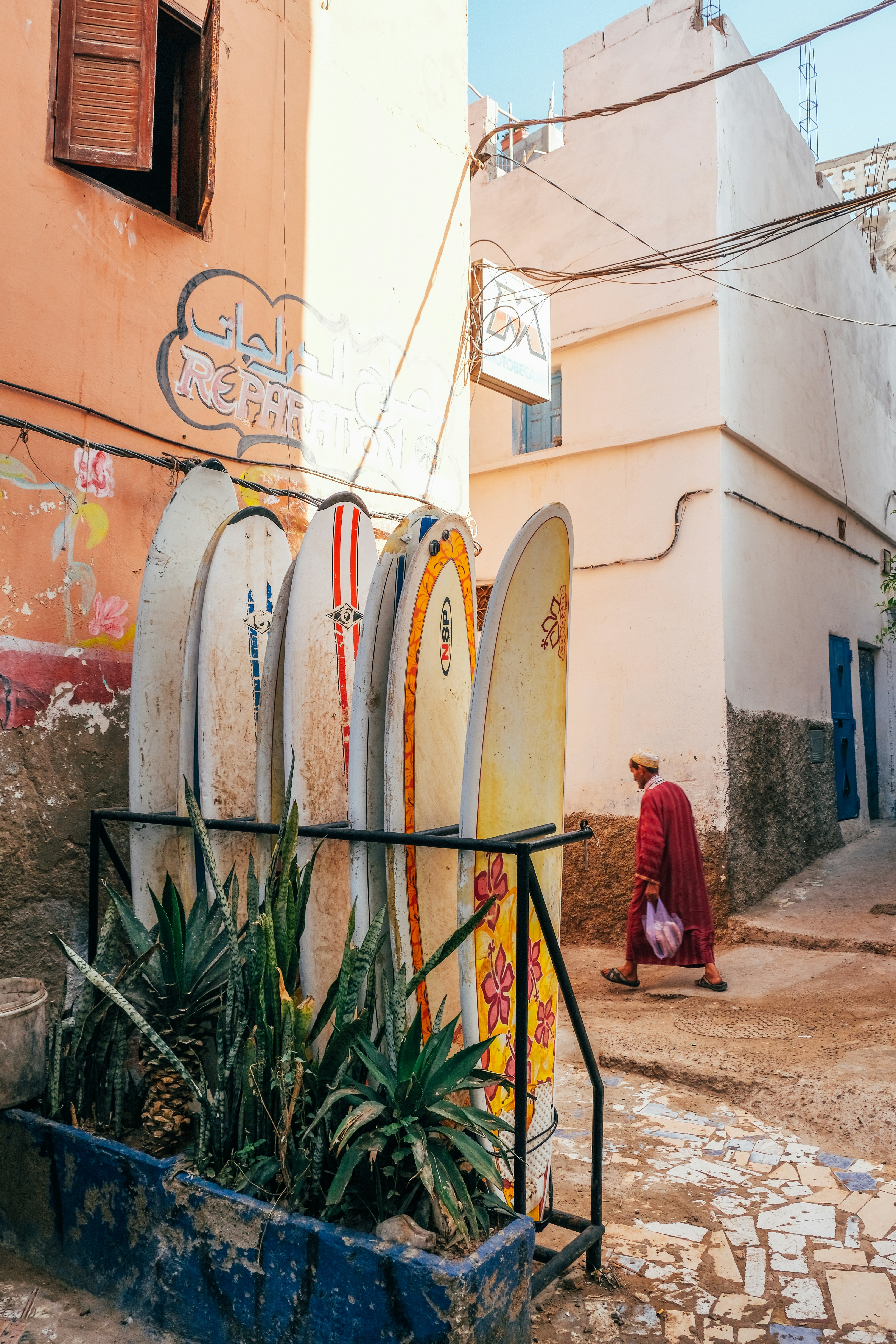 Surfboards lean against a sunlit building, with a man walking by.