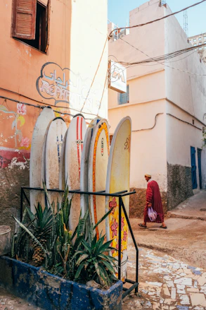 Surfboards lean against a sunlit building, with a man walking by.