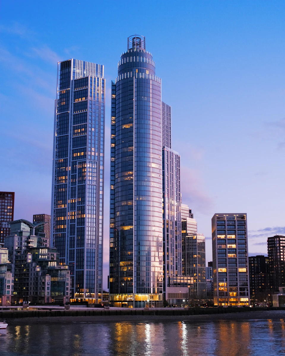 The image depicts a cityscape at dusk, with two tall buildings standing prominently against the sky. The building on the left is a modern skyscraper with a glass facade that reflects the surrounding lights and colors. On the right side of the frame, there's another building with a more traditional design featuring multiple levels and a flat roof. The buildings are situated next to each other, creating an interesting juxtaposition between old and new architectural styles. In the foreground, a body of water can be seen reflecting the city lights, adding depth and dimension to the scene.