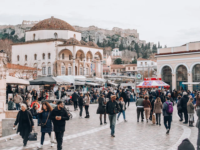 Crowds fill Monastiraki Square in front of the Tzistarakis Mosque, with the Acropolis rising behind.