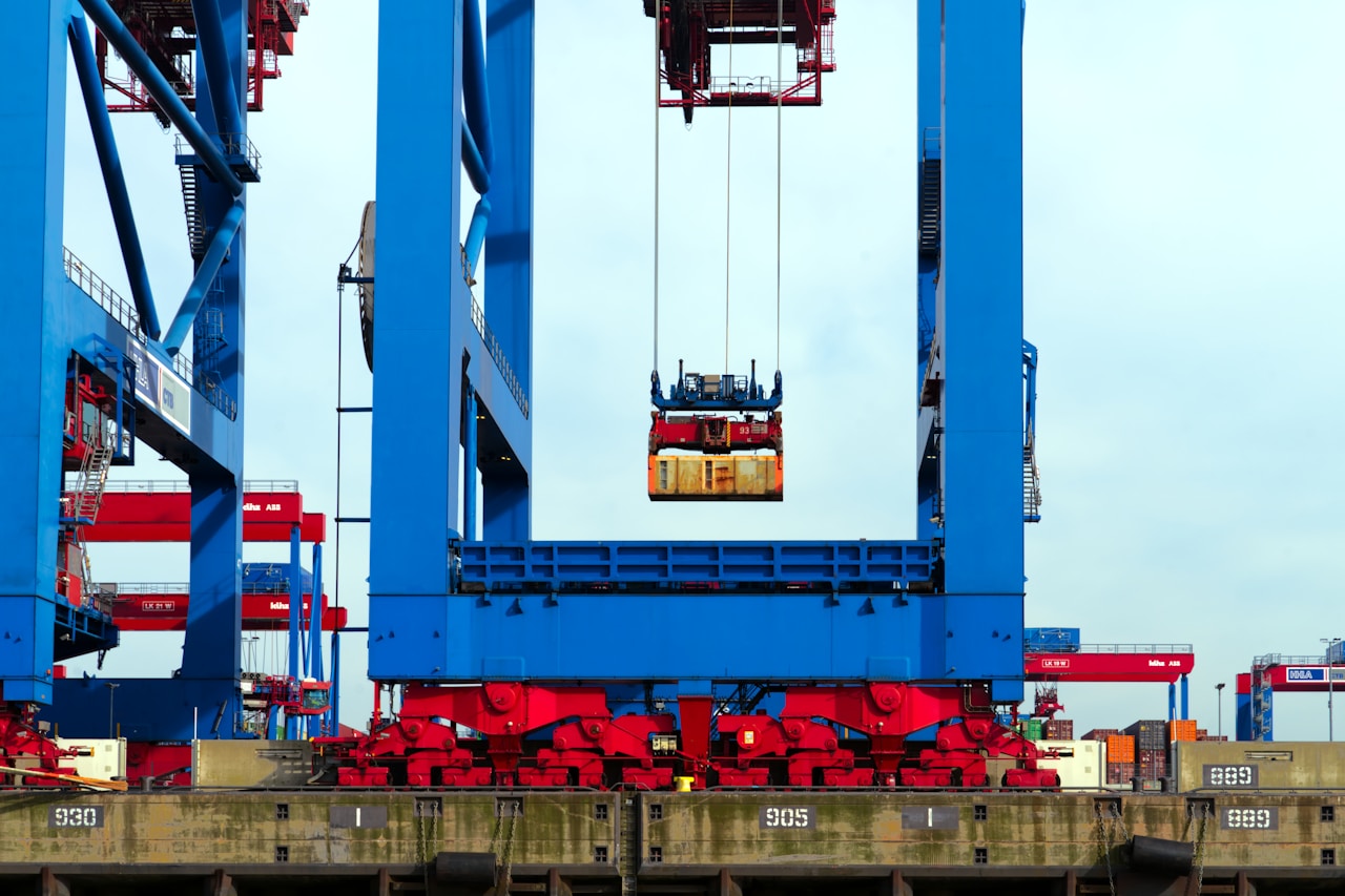 A quay crane carrying a container in mid-air, with blue gantry cranes behind it