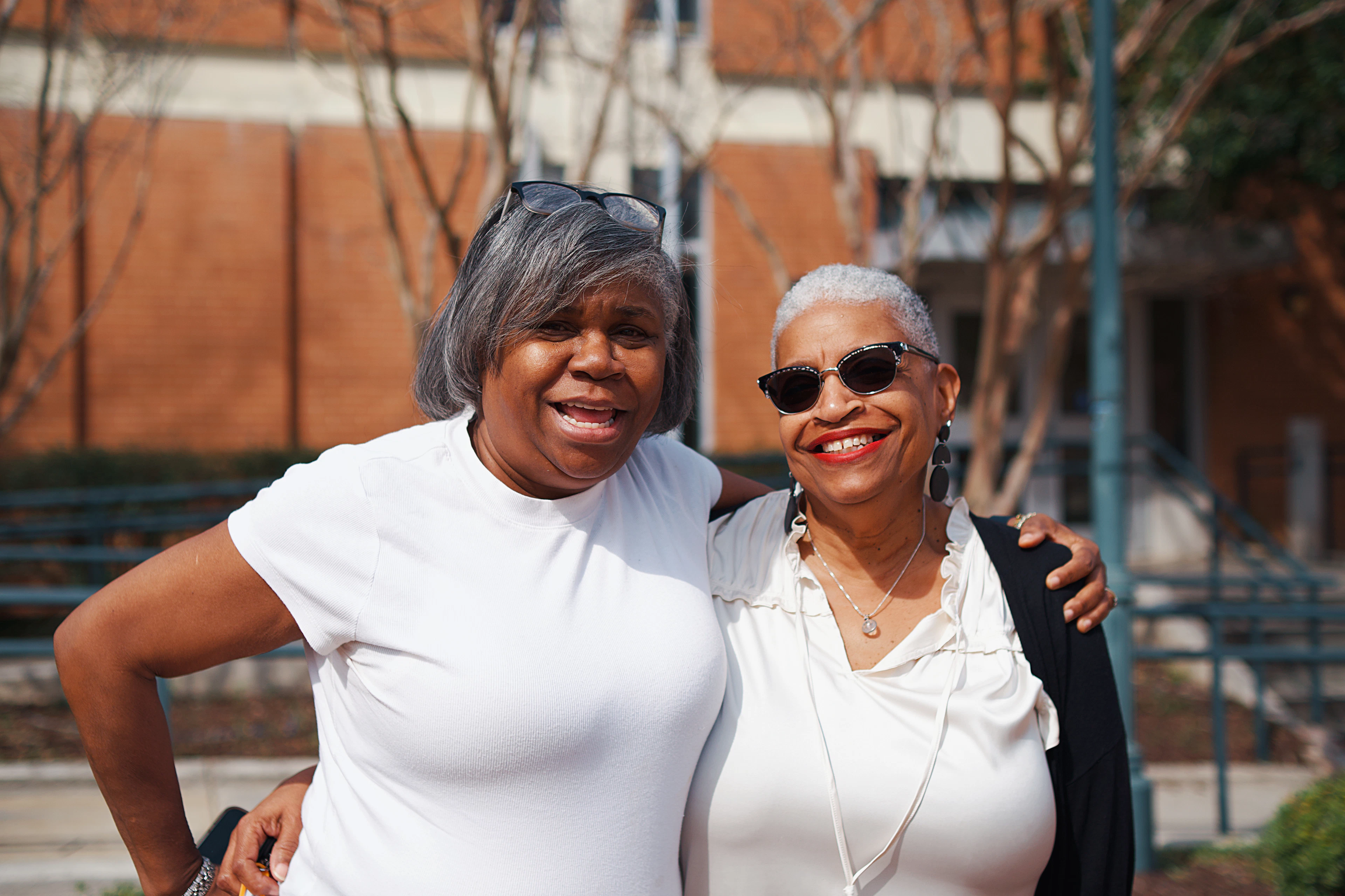 Two women standing arm in arm outdoors, both smiling at the camera