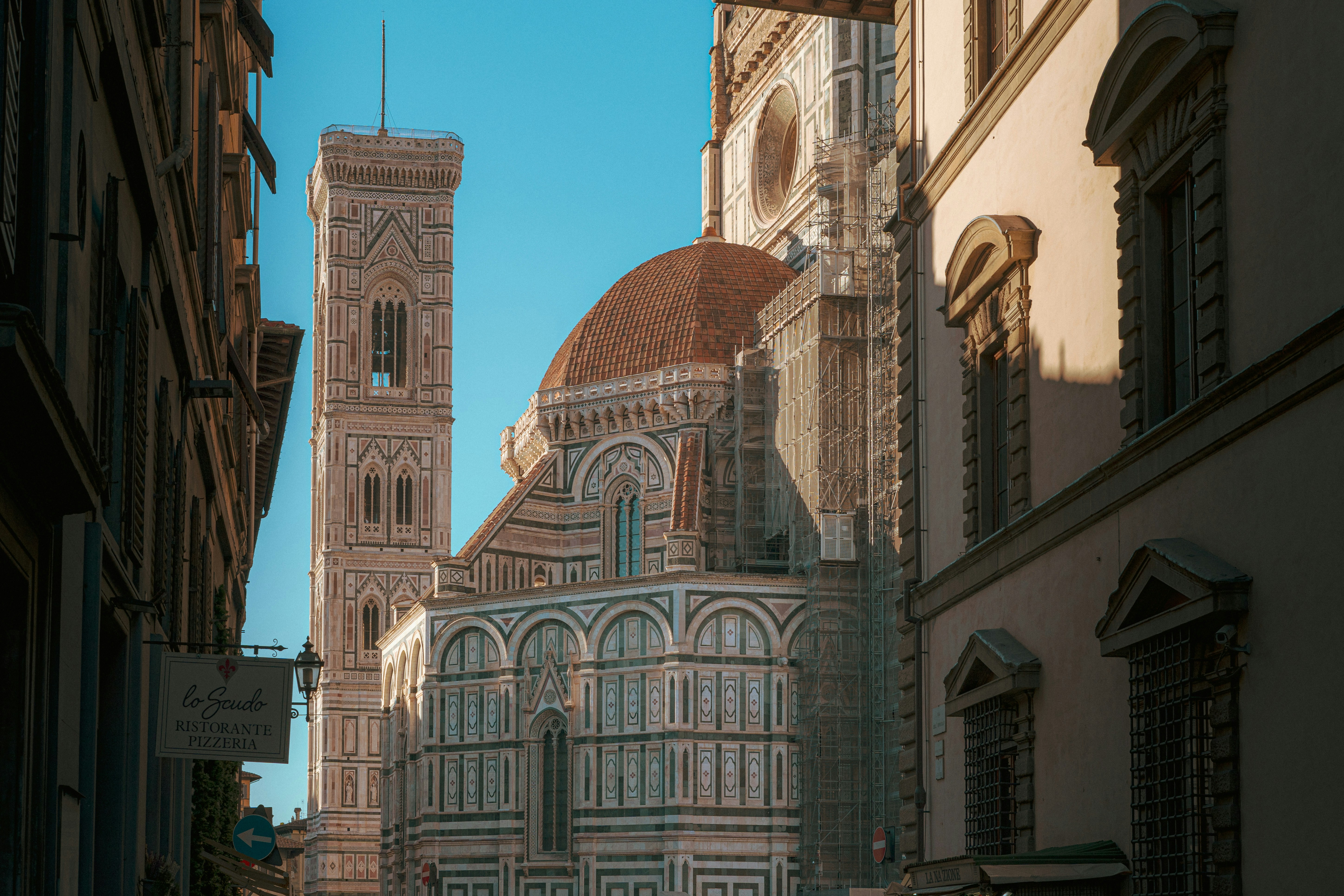 Majestätische Ansicht der Kathedrale von Florenz (Cattedrale di Santa Maria del Fiore) mit der ikonischen roten Kuppel von Brunelleschi und dem Glockenturm von Giotto, eingerahmt von historischen Gebäuden in warmem Sonnenlicht. Perfekte Architekturaufnahme für Reise- und Italien-Liebhaber.