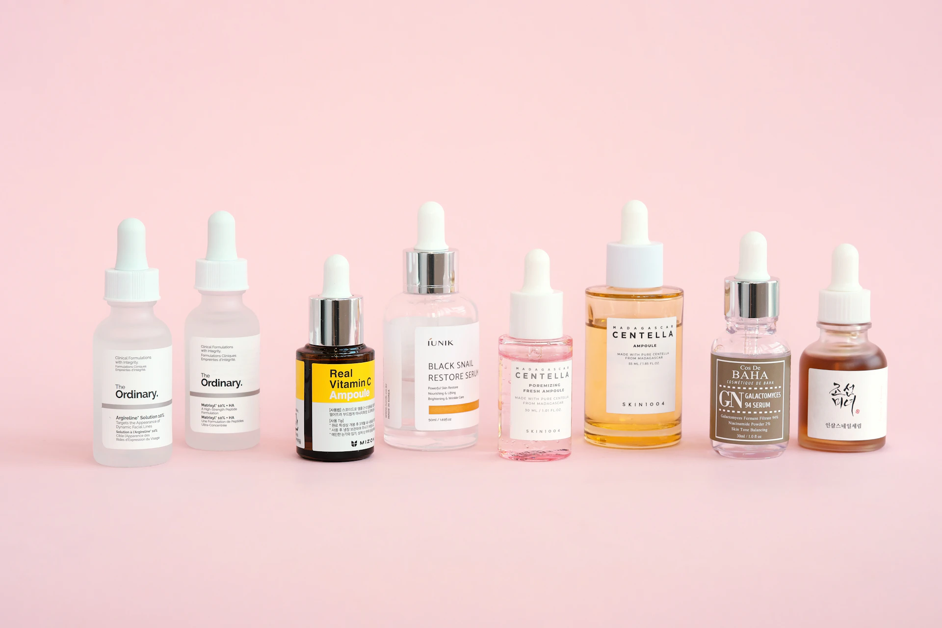 Skincare bottles are displayed on a pastel pink background.
