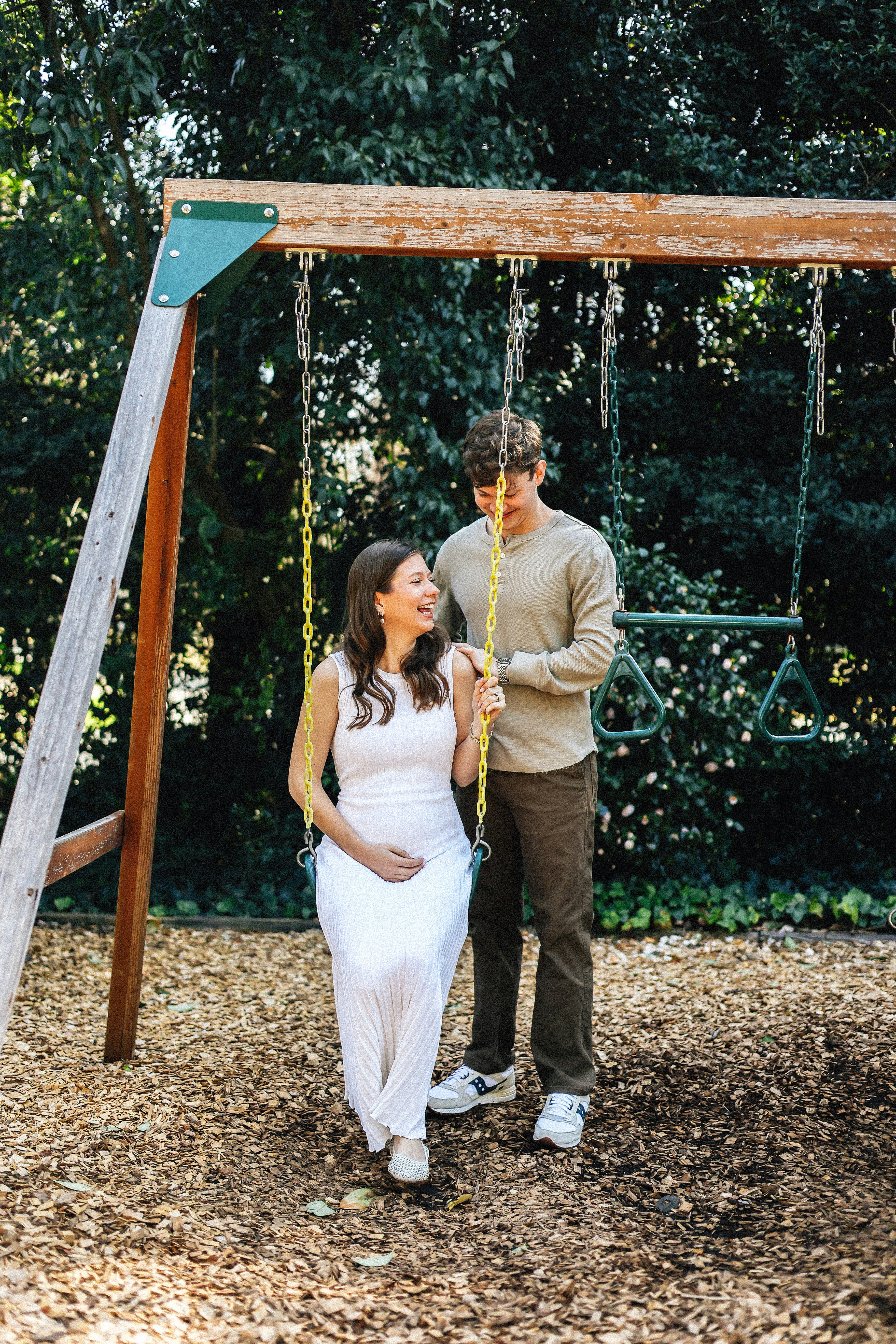 Pregnant couple poses together on a swing set.