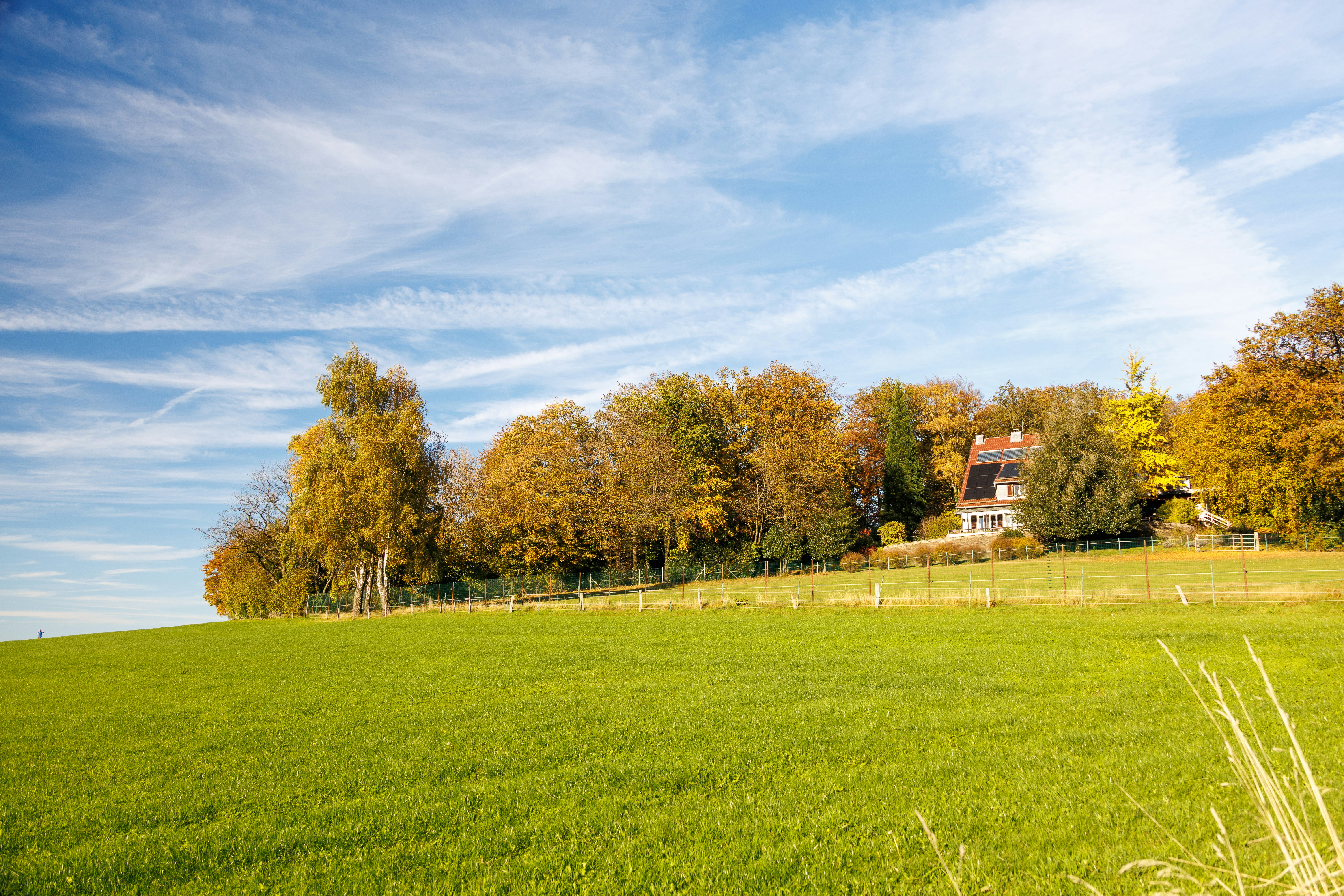 A house sits behind a green field and autumn trees.