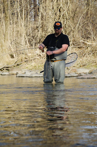An angler in chest waders standing thigh-deep in flat water, fly rod in hand and a landing net on the belt, working line through the current