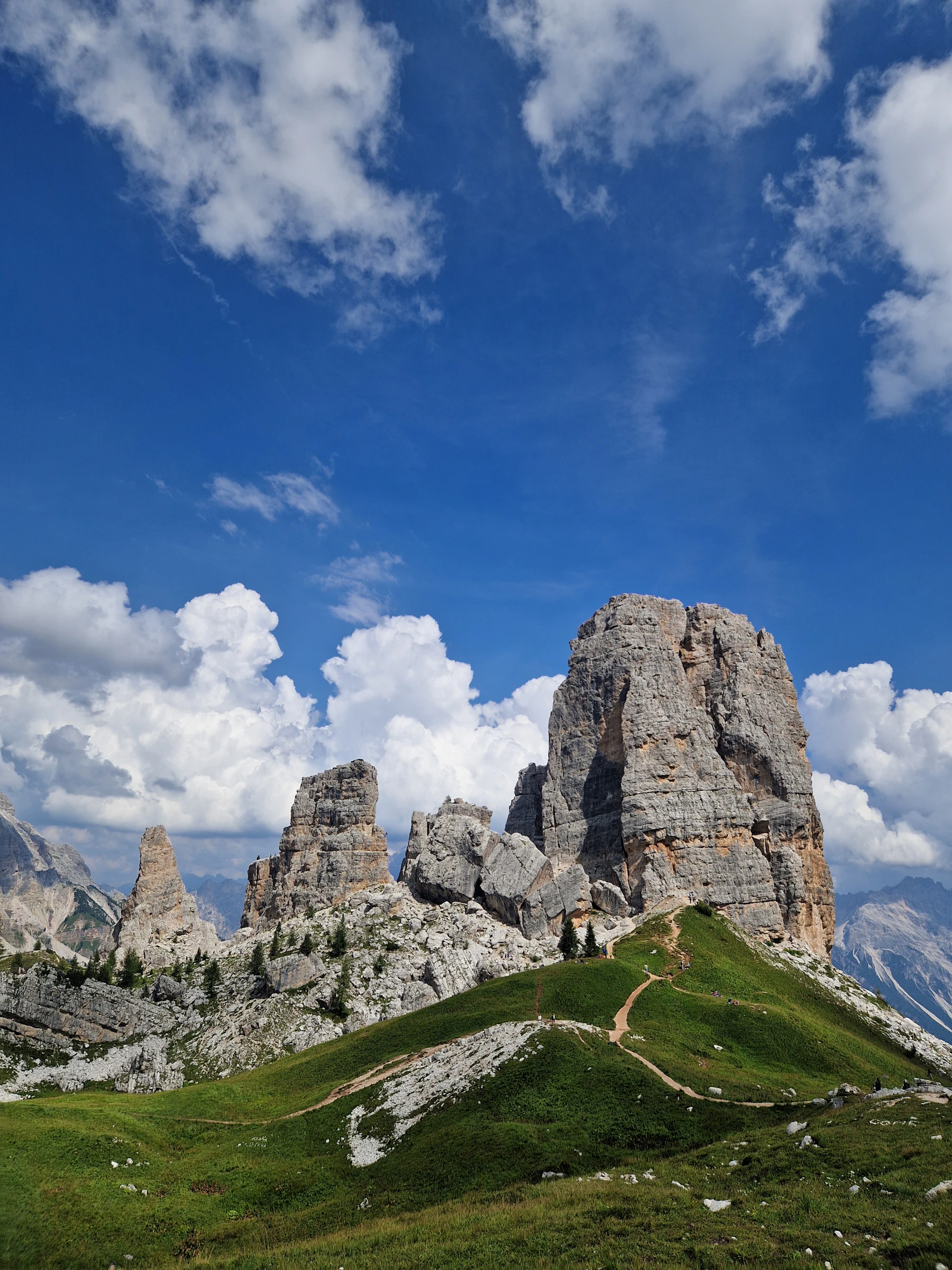 Photograph of A tall Cinque Torri rock formation rises above pale paths beneath a wide blue sky.