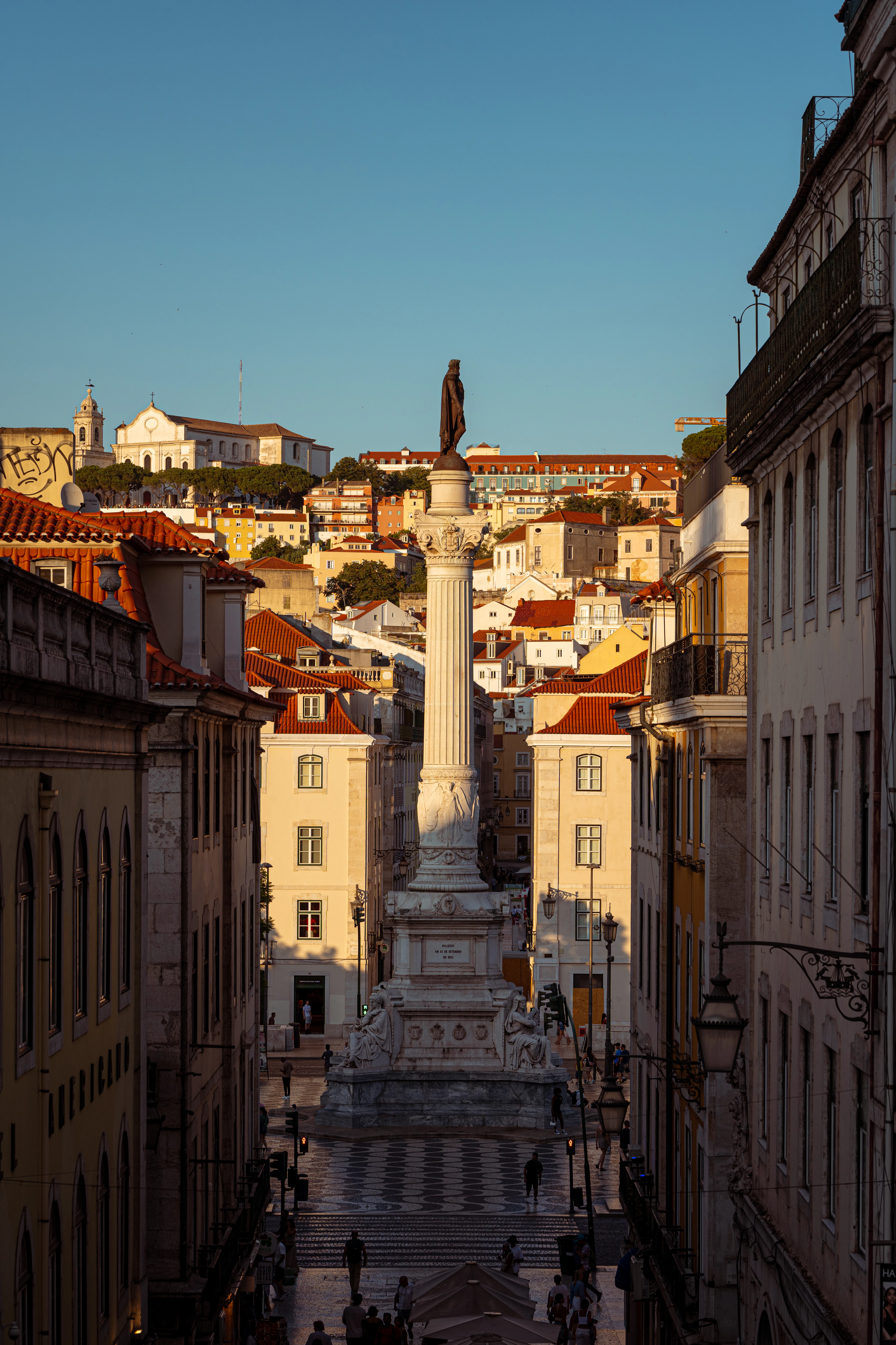 The column of Dom Pedro IV rising over Rossio Square in Lisbon at golden hour, framed between tall pastel townhouses with the hillside of Graca and its white church behind