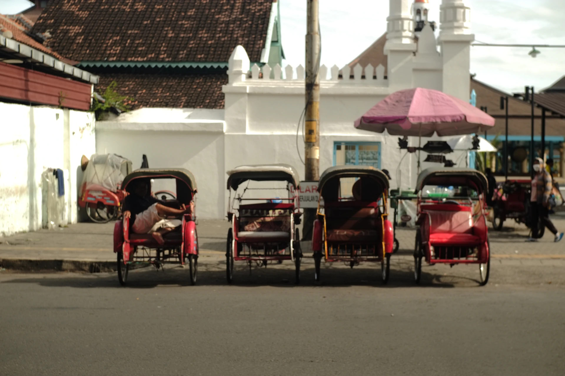 Rickshaws line a street near a white building.
