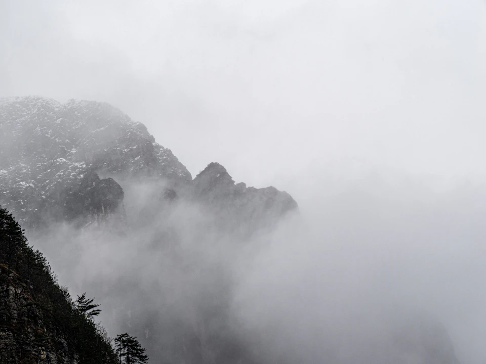 Foggy mountain range with mist-shrouded peaks and snow-covered terrain in a hazy gray sky