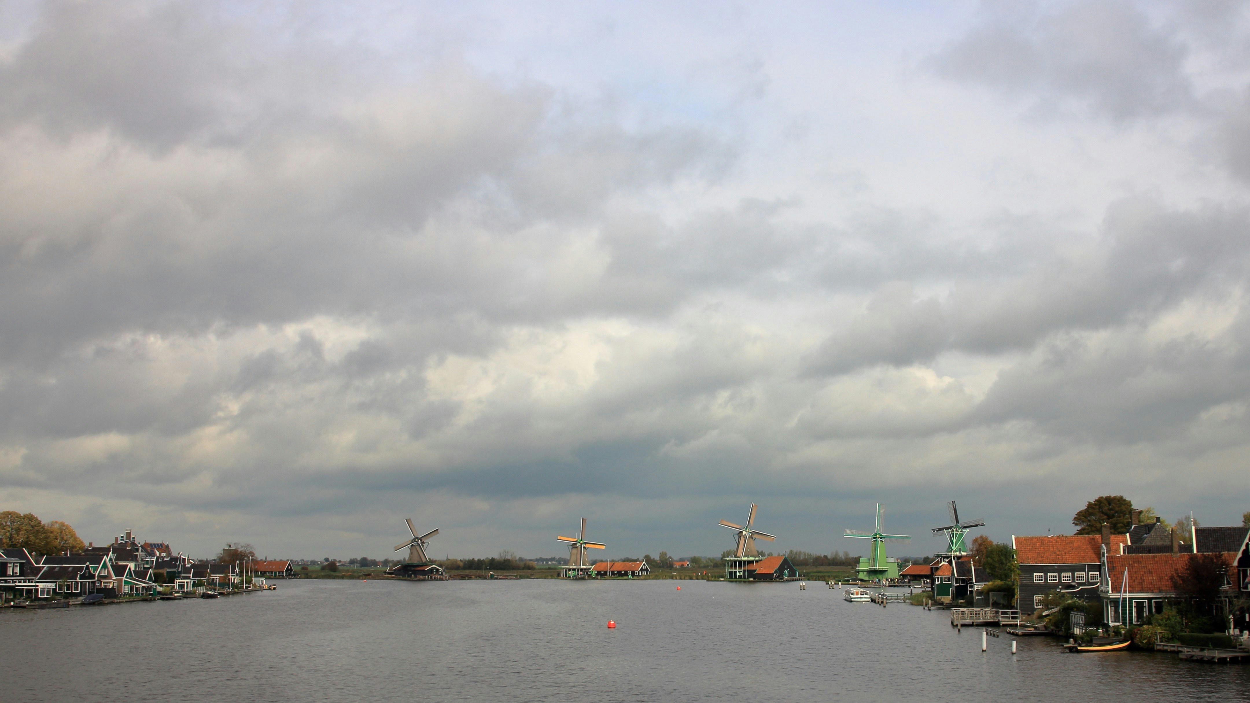Windmühlen und Häuser säumen einen Fluss unter bewölktem Himmel.