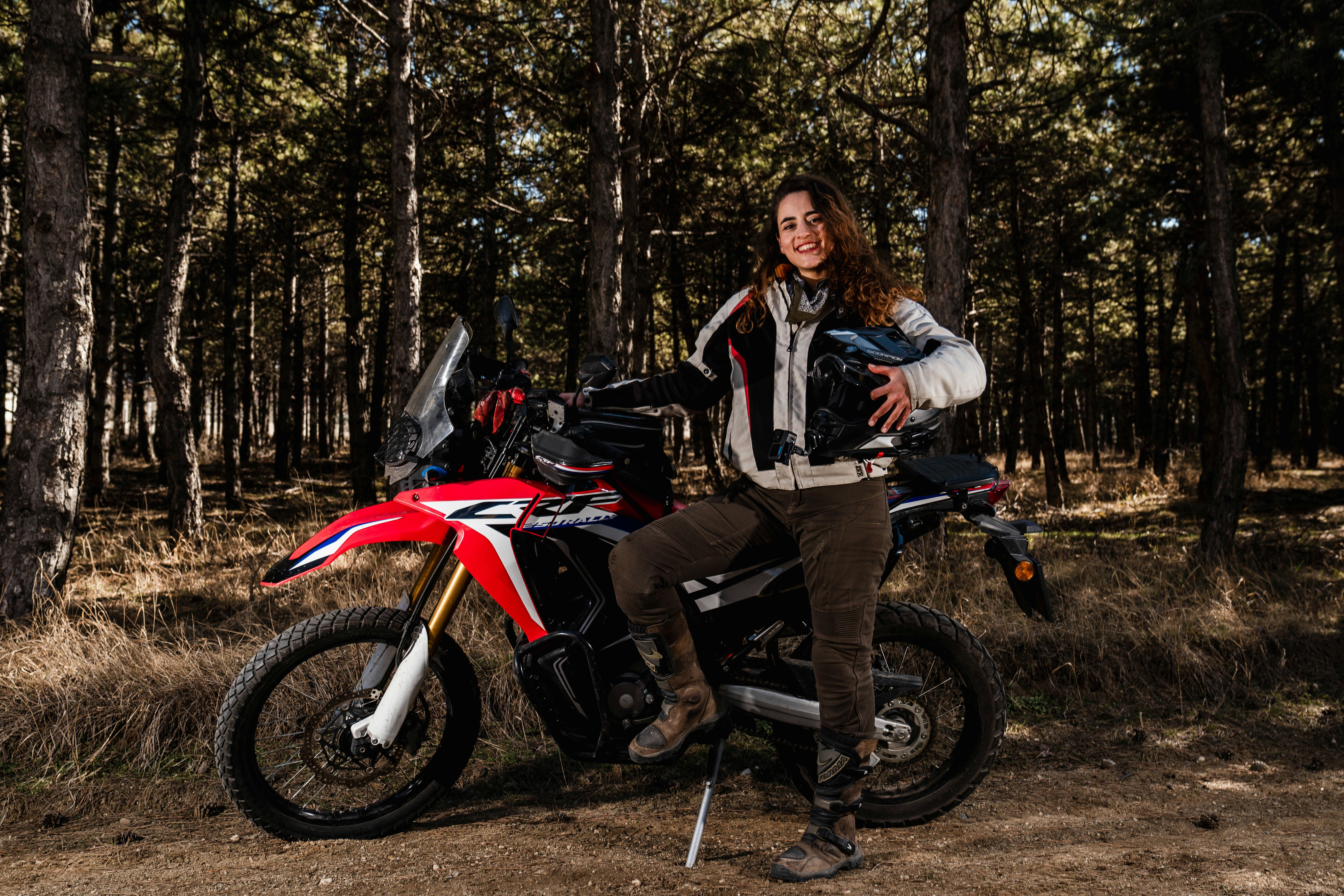 Woman poses next to her motorcycle in woods.