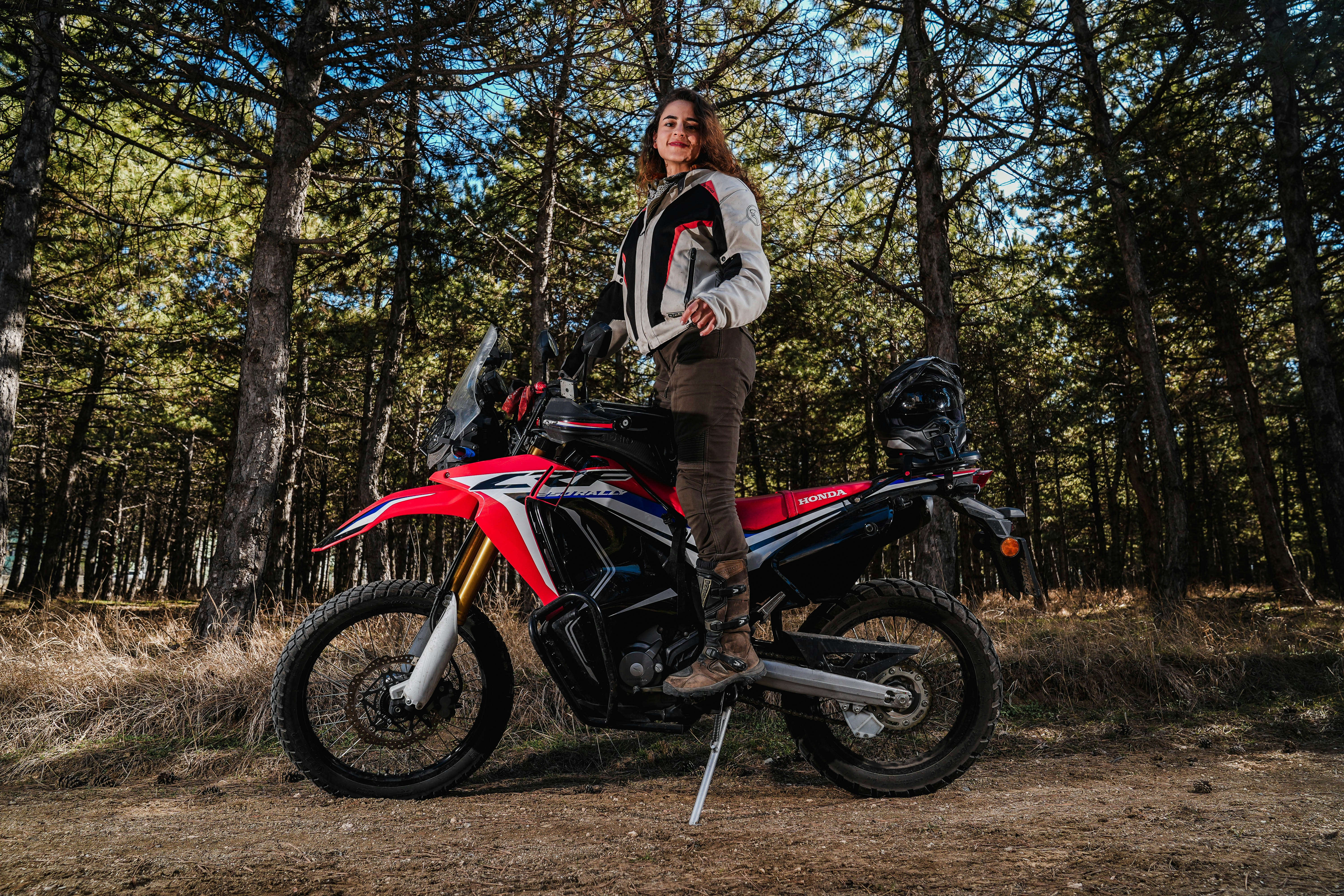 Woman poses with her red motorcycle in the woods.