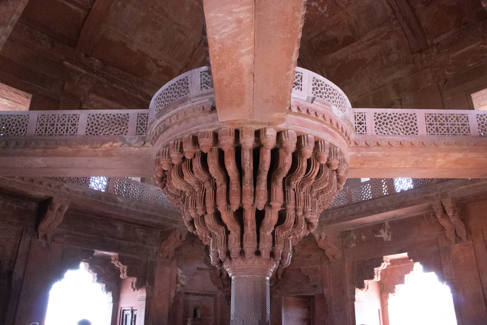 The image shows a large, ornate stone structure with intricate carvings and designs. The central feature is a tall, cylindrical column adorned with numerous carved patterns on its surface. The column is supported by four sturdy columns at each corner, creating an impressive architectural design. The walls surrounding the column are also decorated with similar carvings, adding to the overall grandeur of the structure. The image is taken from a low angle, emphasizing the height and scale of the column and the intricate details of the carvings.
